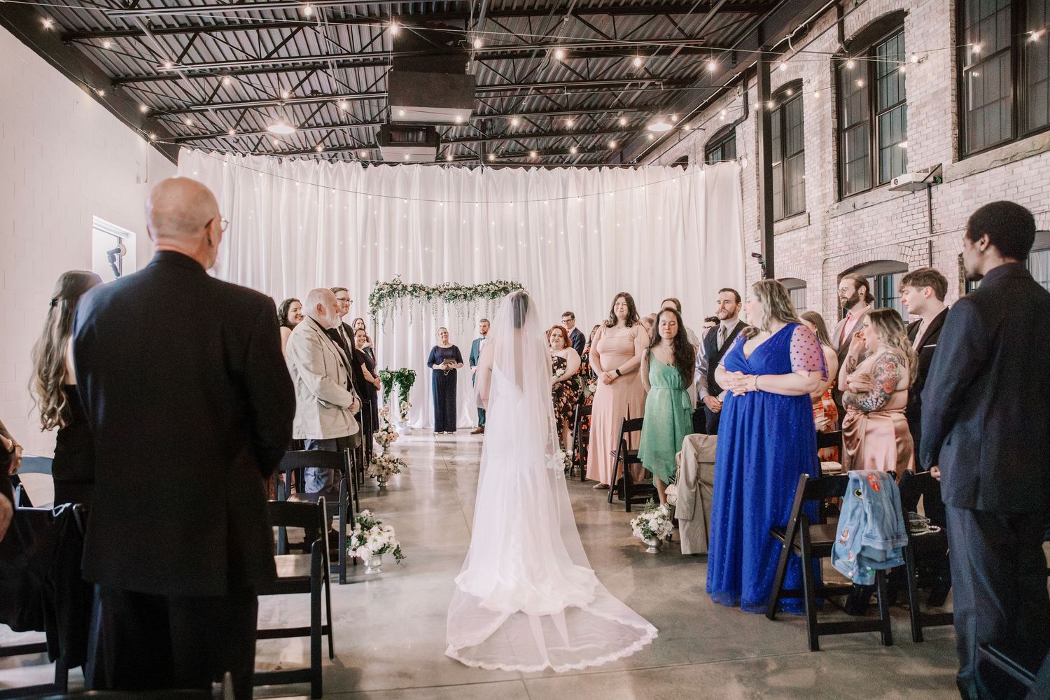 Wedding ceremony setup with white draping and guests seated in industrial venue space.