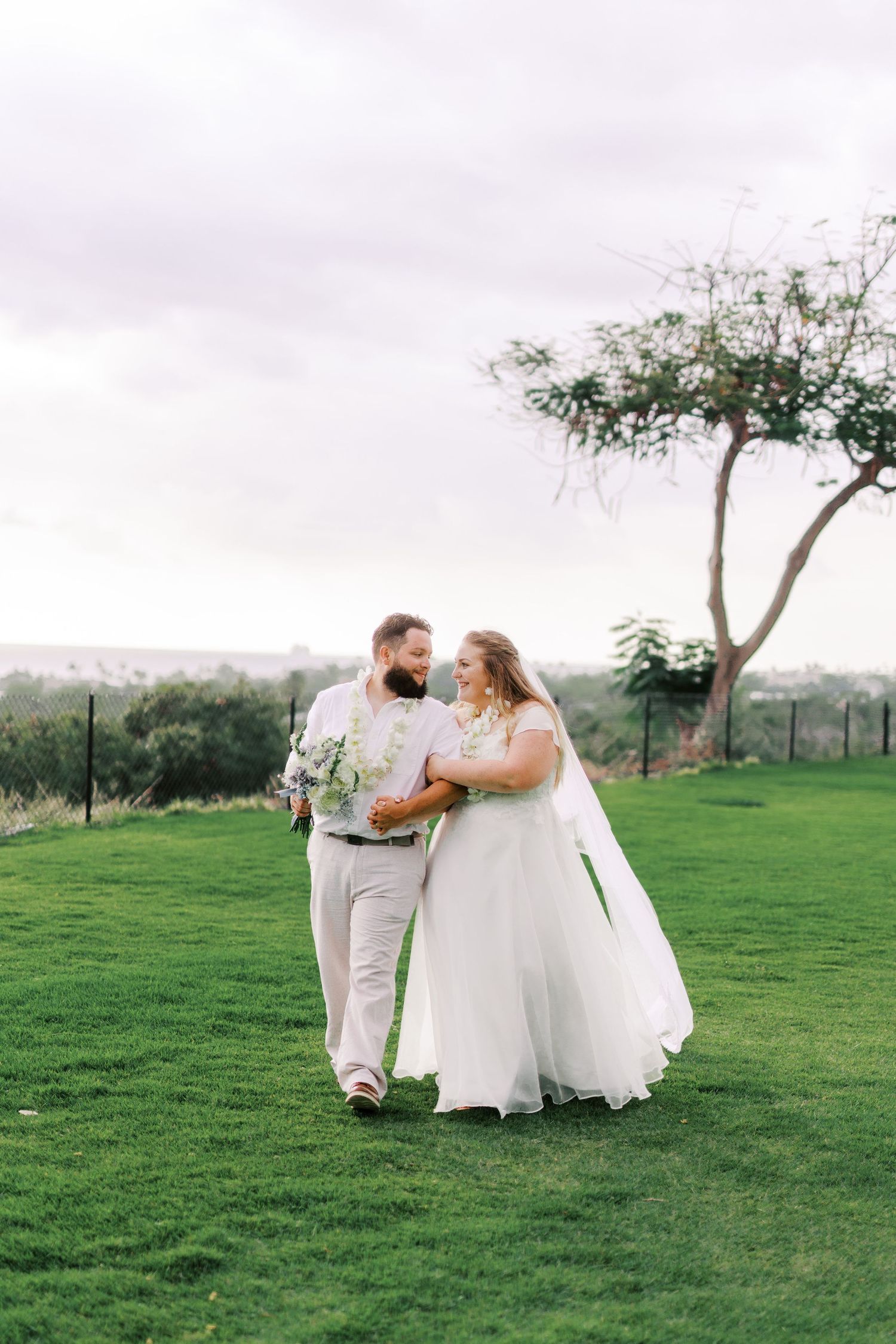 A newlywed couple walks together on a lush green lawn with a scenic backdrop.