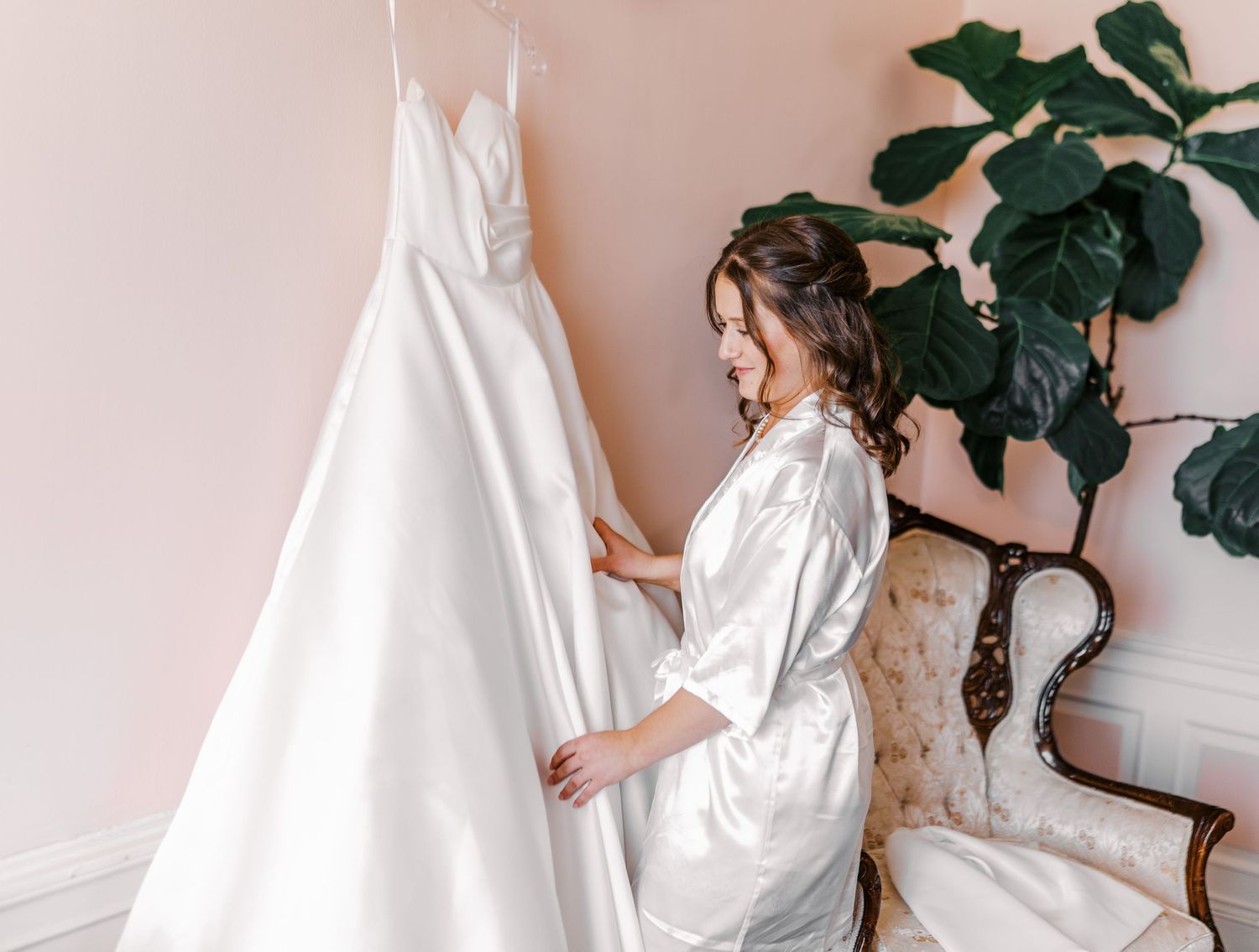 Someone in a white silk robe admires an elegant white wedding dress hanging against a pink wall with a fiddle leaf fig plant.