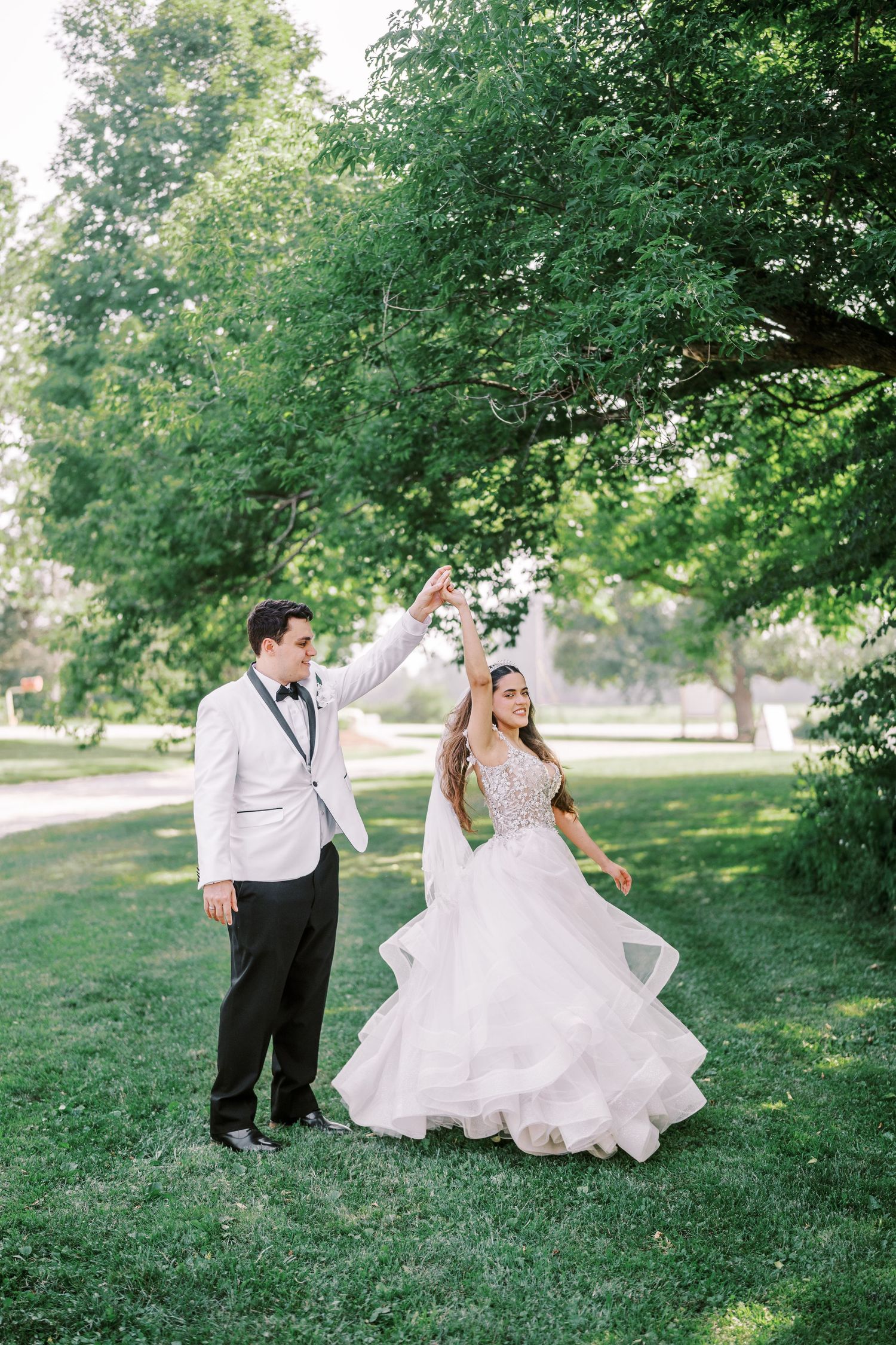 A couple share a romantic dance moment on a lush green lawn beneath towering trees on their wedding day.