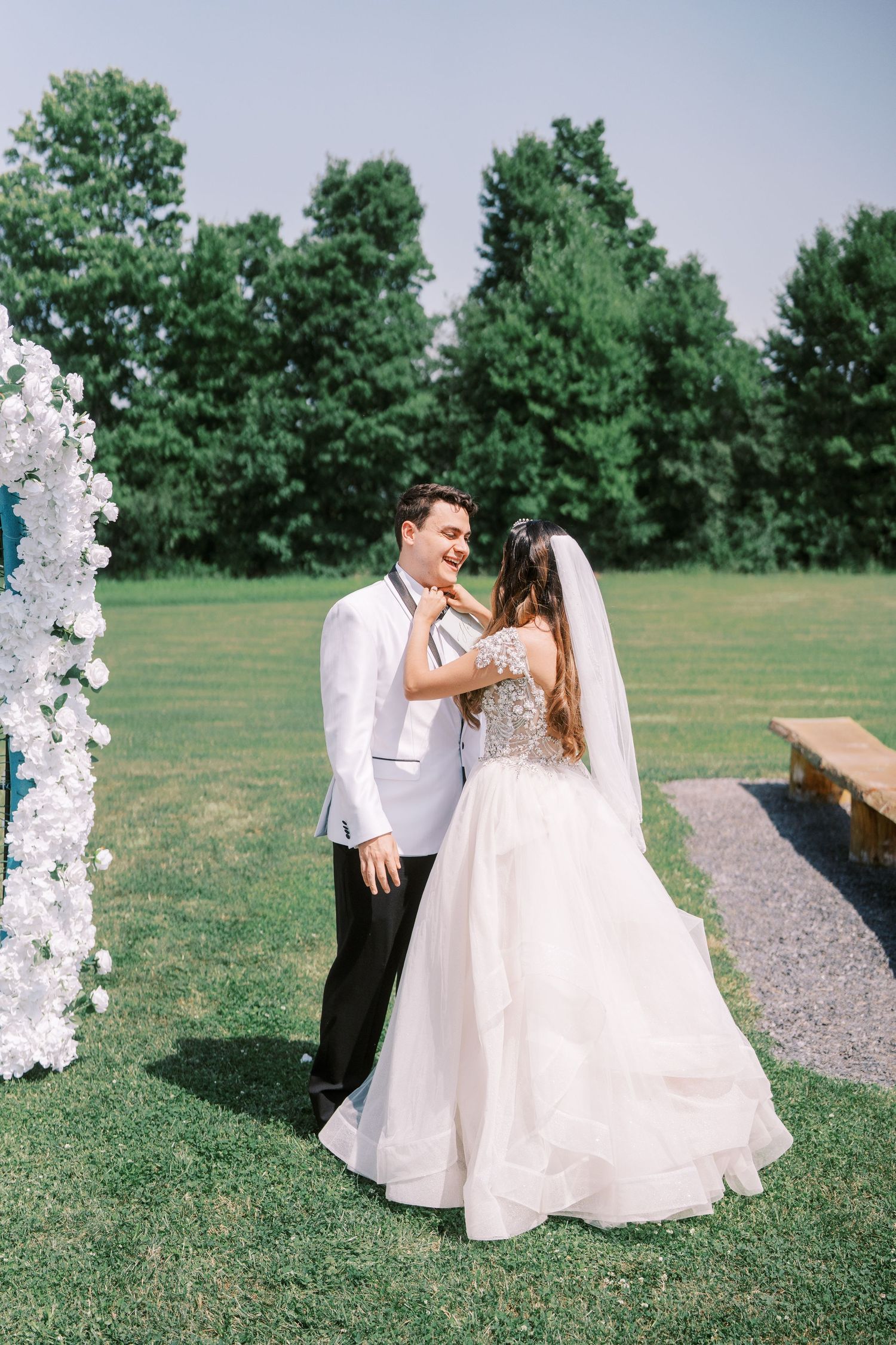 Wedding couple shares a joyful moment on a paved path surrounded by lush green grass and trees on a sunny day.