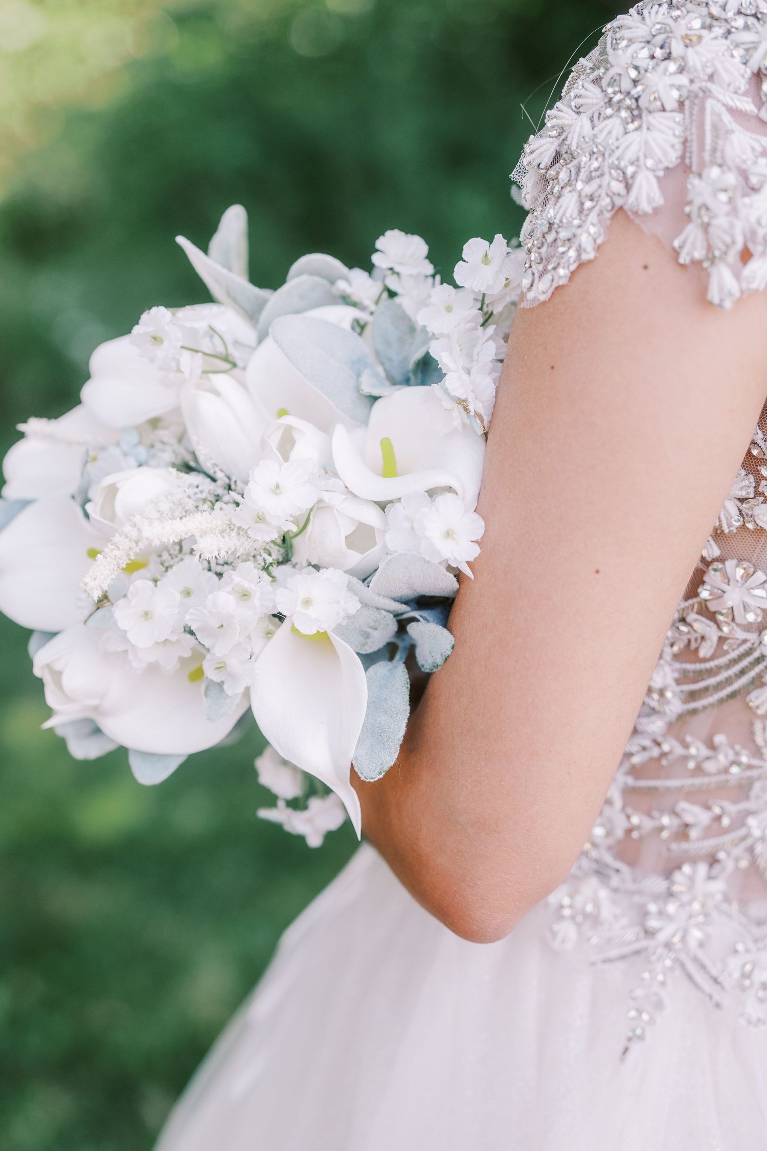 A delicate white bridal bouquet with orchids and lilies is held against an intricately beaded lace wedding gown.