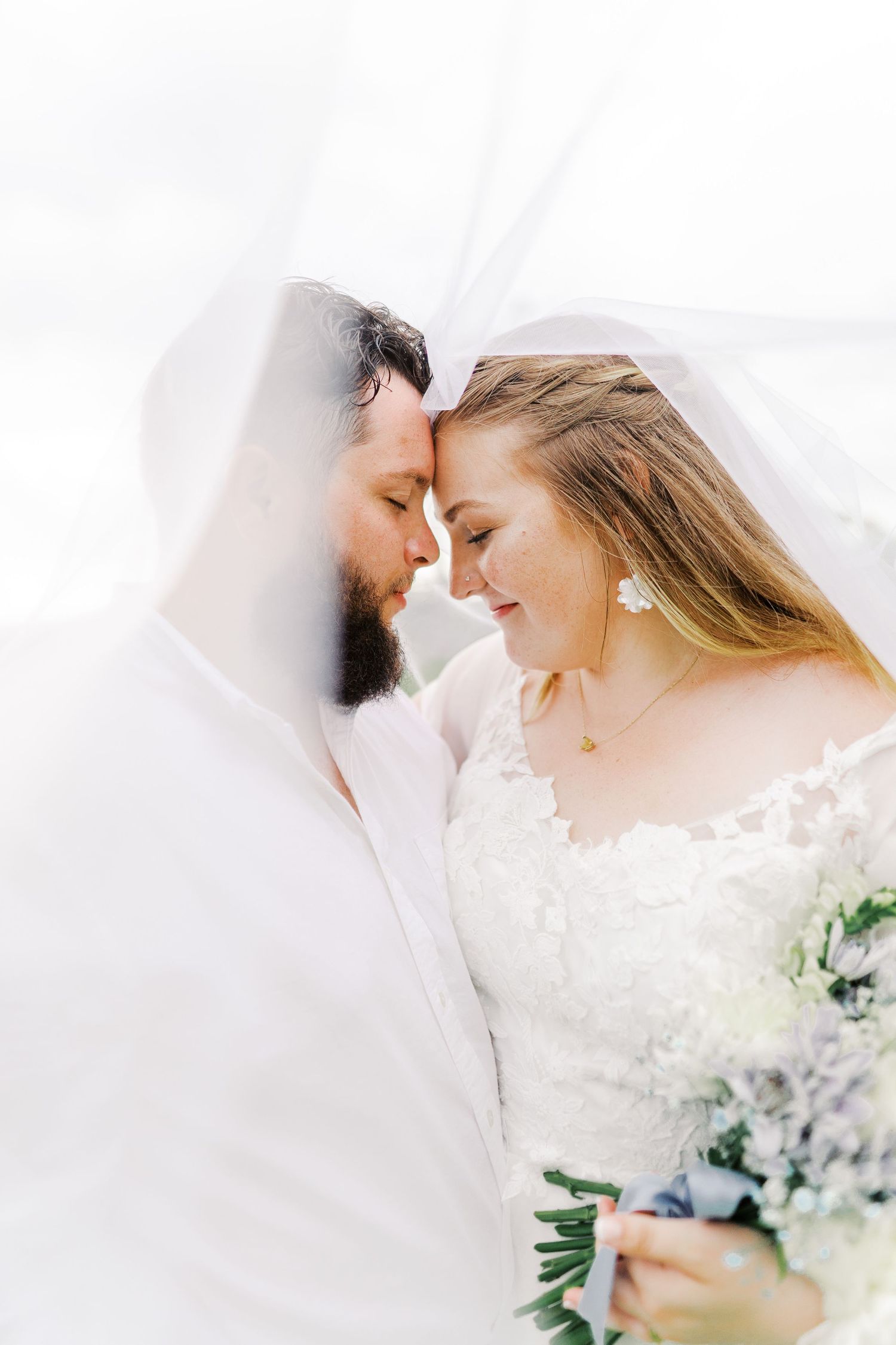 A tender moment between newlyweds under a veil with soft lighting and a white and blue floral bouquet.