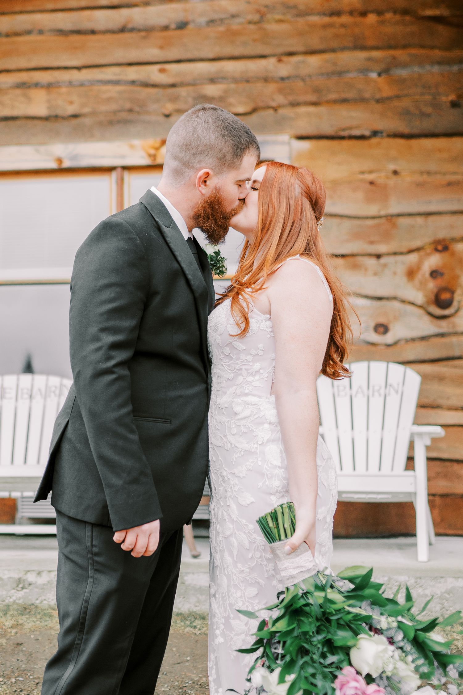 Romantic moment between newlyweds sharing a kiss against rustic wooden cabin backdrop.