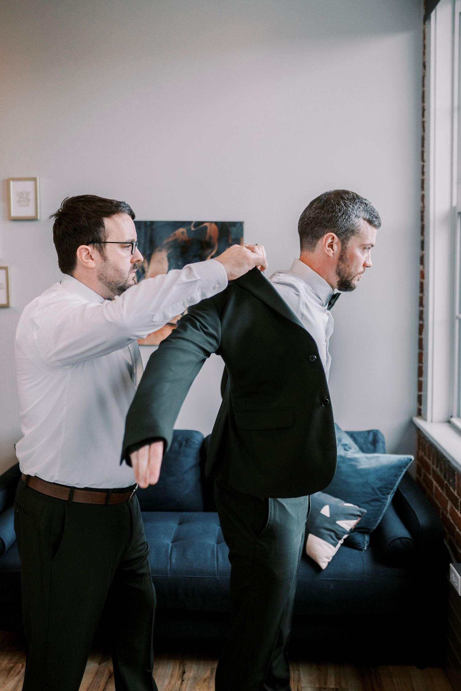 Two men in formal attire helping with getting dressed, with one assisting the other in putting on a jacket near a blue couch.