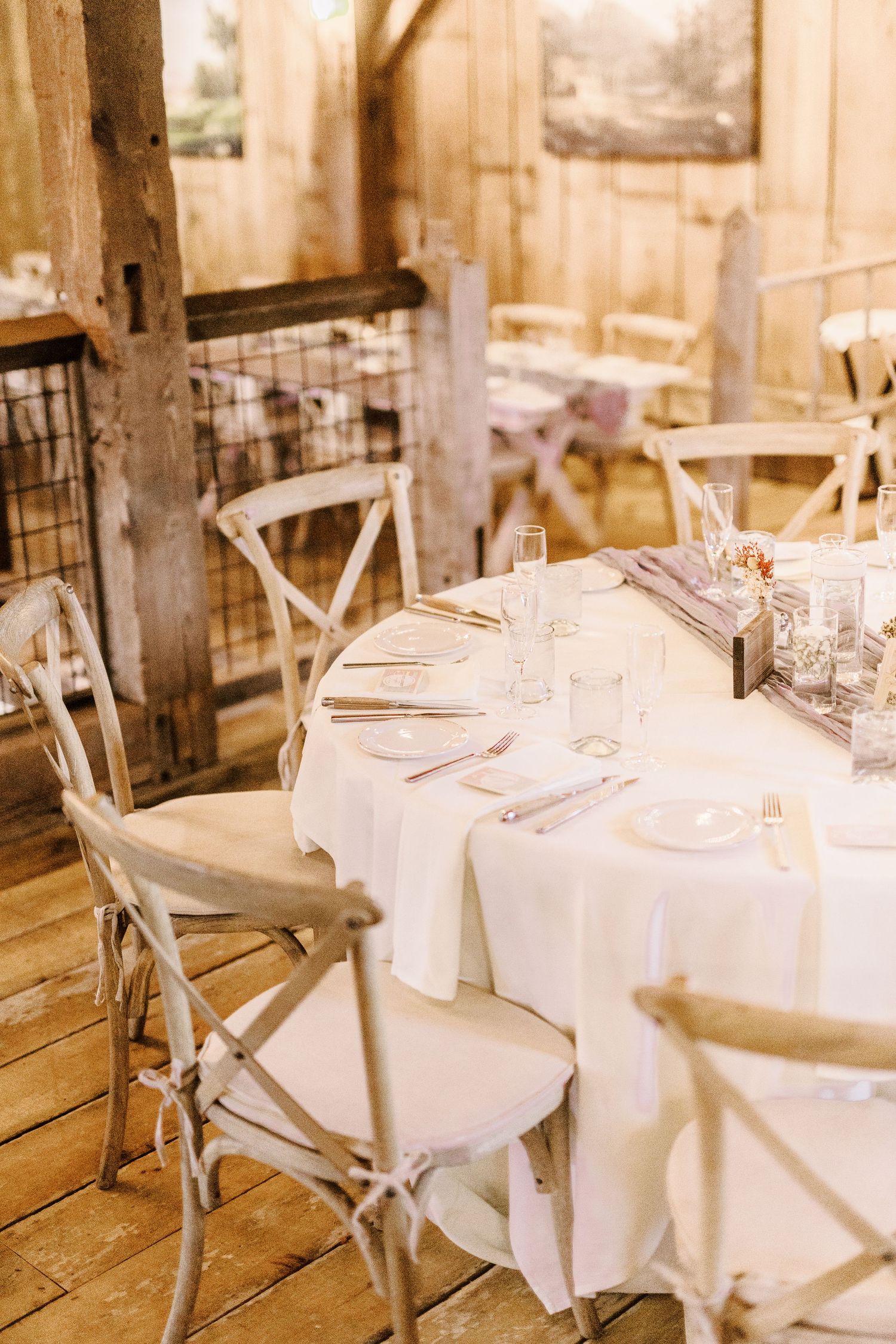 Elegant rustic wedding reception table with white linens and cross-back chairs in a barn venue.