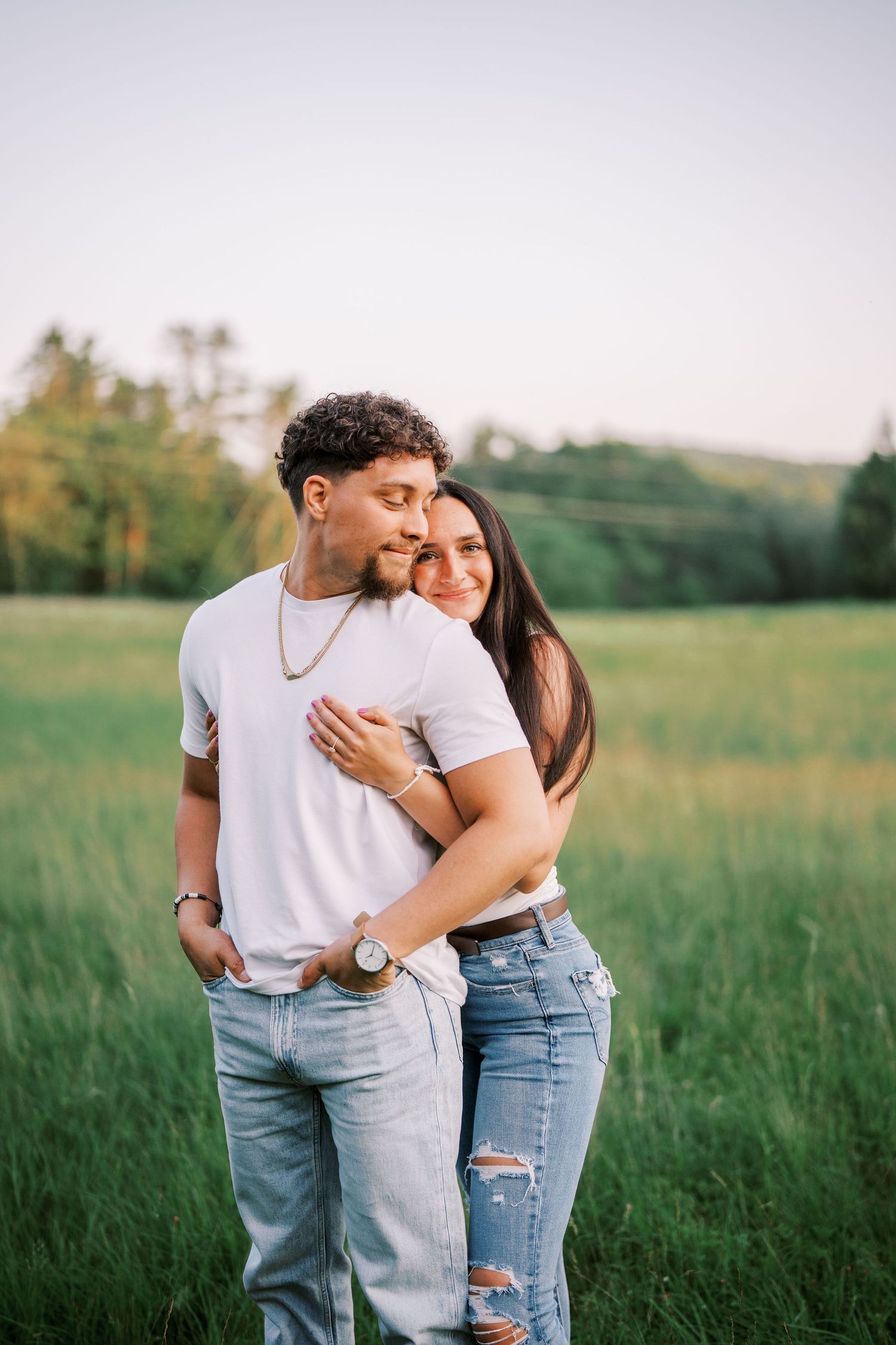 Two people in matching white tops and denim embrace affectionately in a sunlit meadow with tall grass.