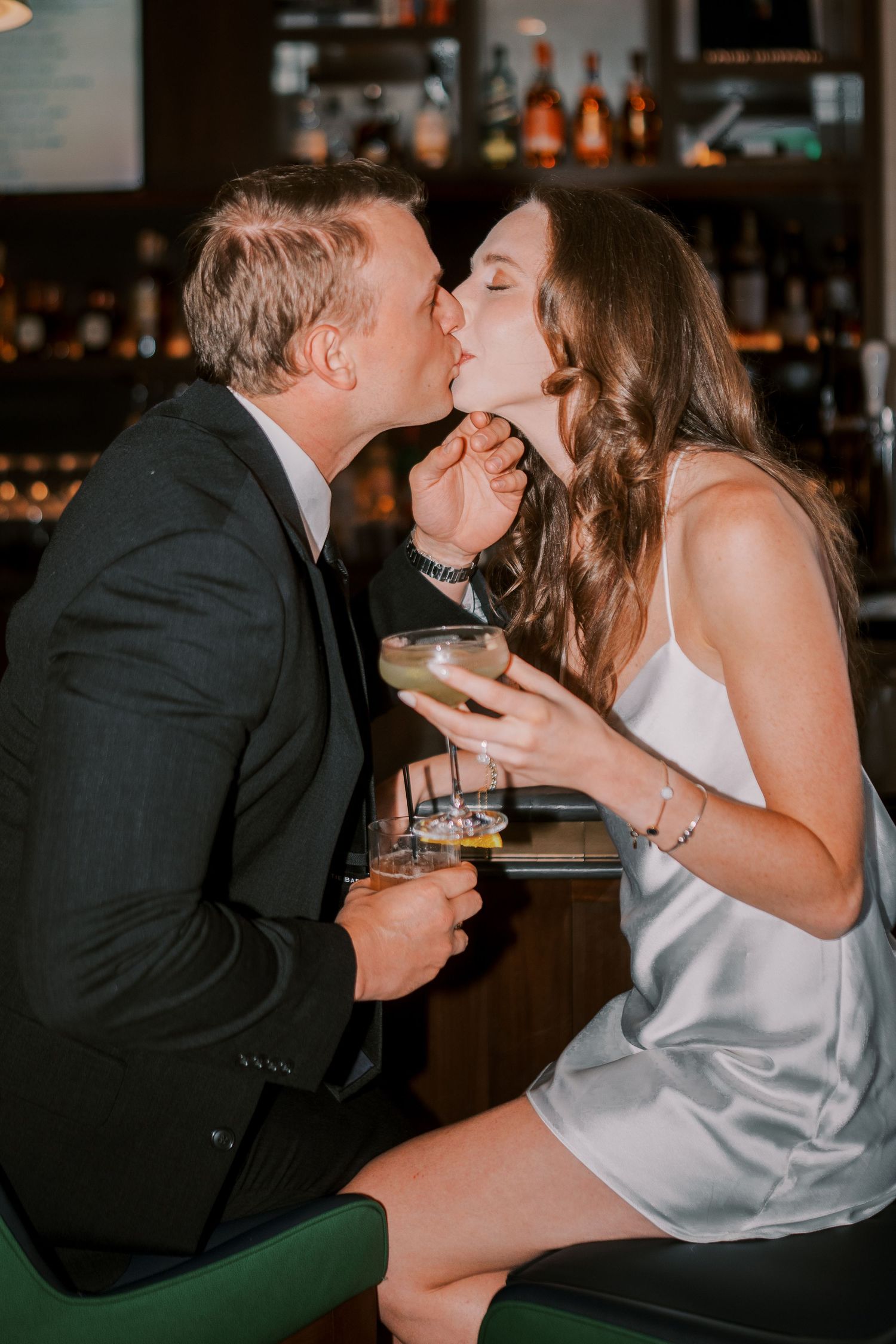 Romantic couple sharing a kiss at a bar while holding cocktail drinks in a dimly lit setting.