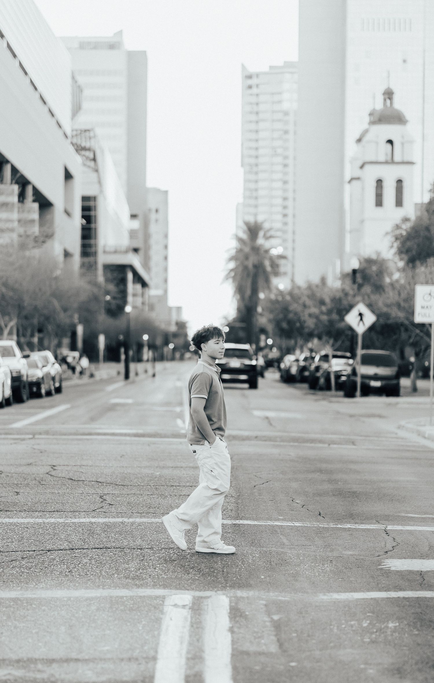 Urban street scene with person crossing road between parked cars and tall buildings in black and white photography.