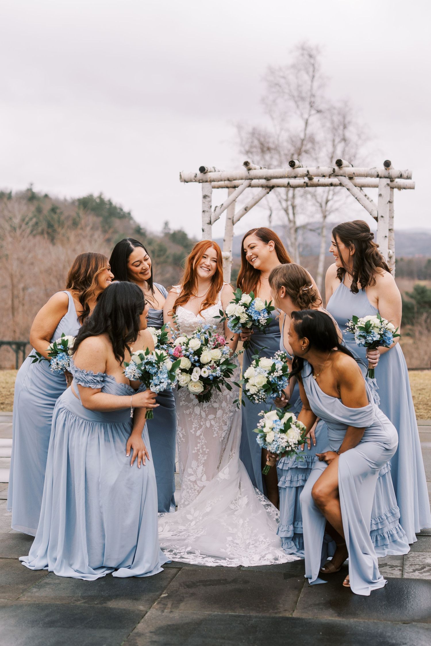 Bridesmaids in flowing light blue dresses gather around bride under rustic wooden arch with bouquets.