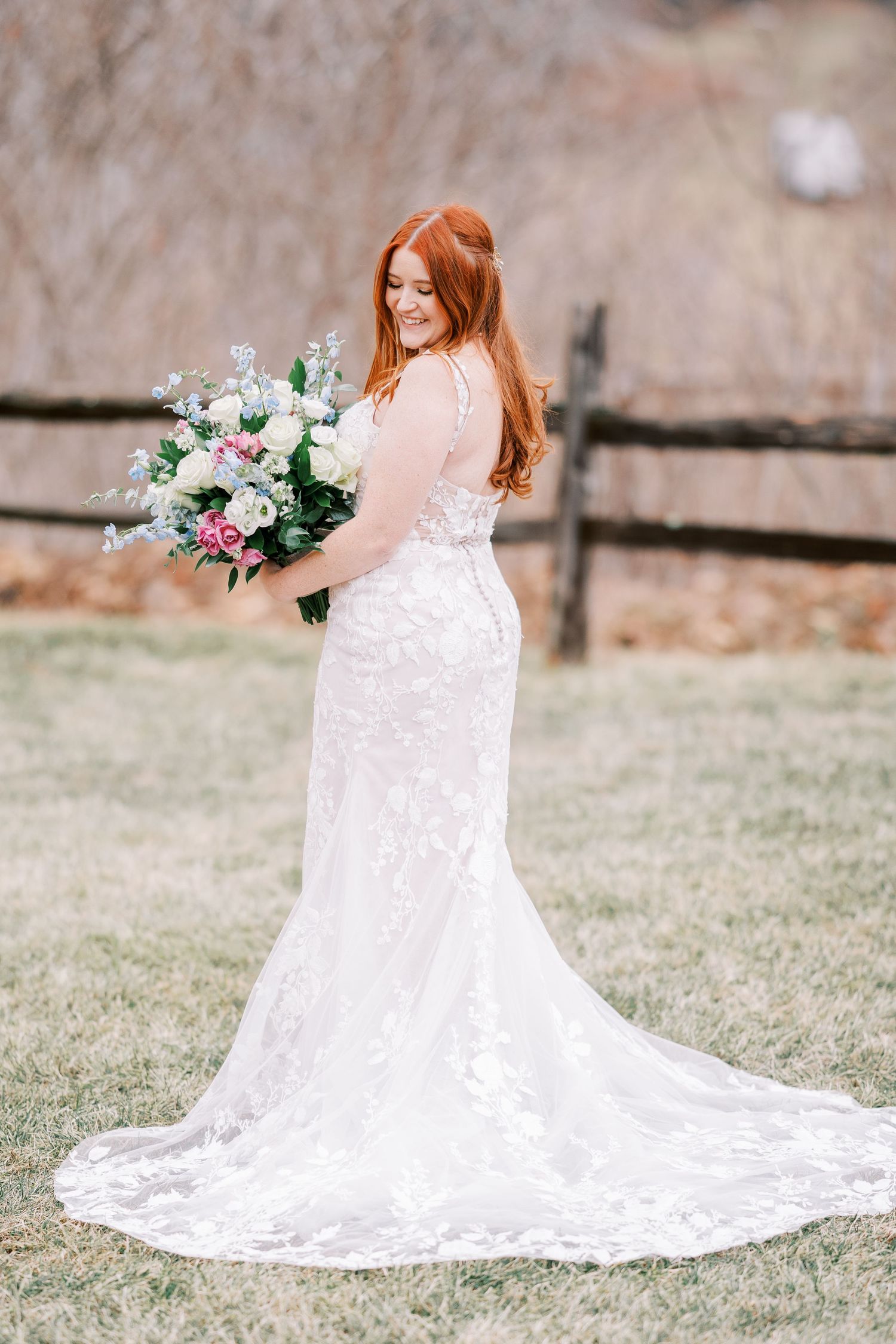 Bride in lace wedding dress with long train poses with bouquet in outdoor rustic setting.