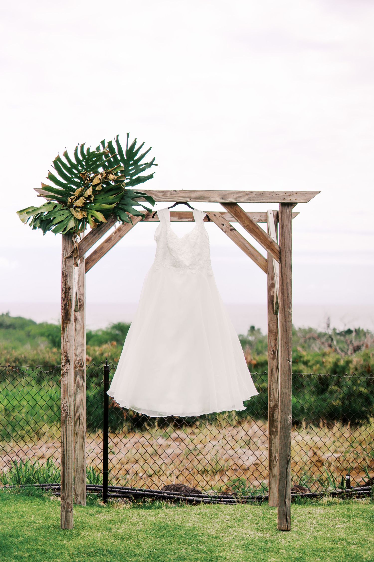 A white wedding dress hangs from a rustic wooden arch decorated with tropical greenery.