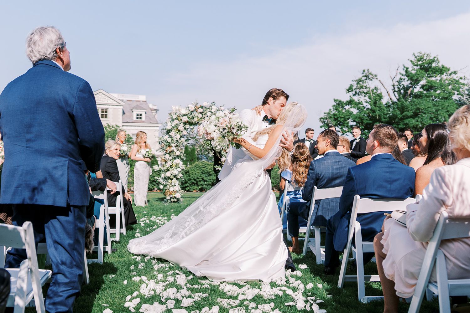 An outdoor garden wedding ceremony with white floral petals lining the aisle and guests seated in white chairs.
