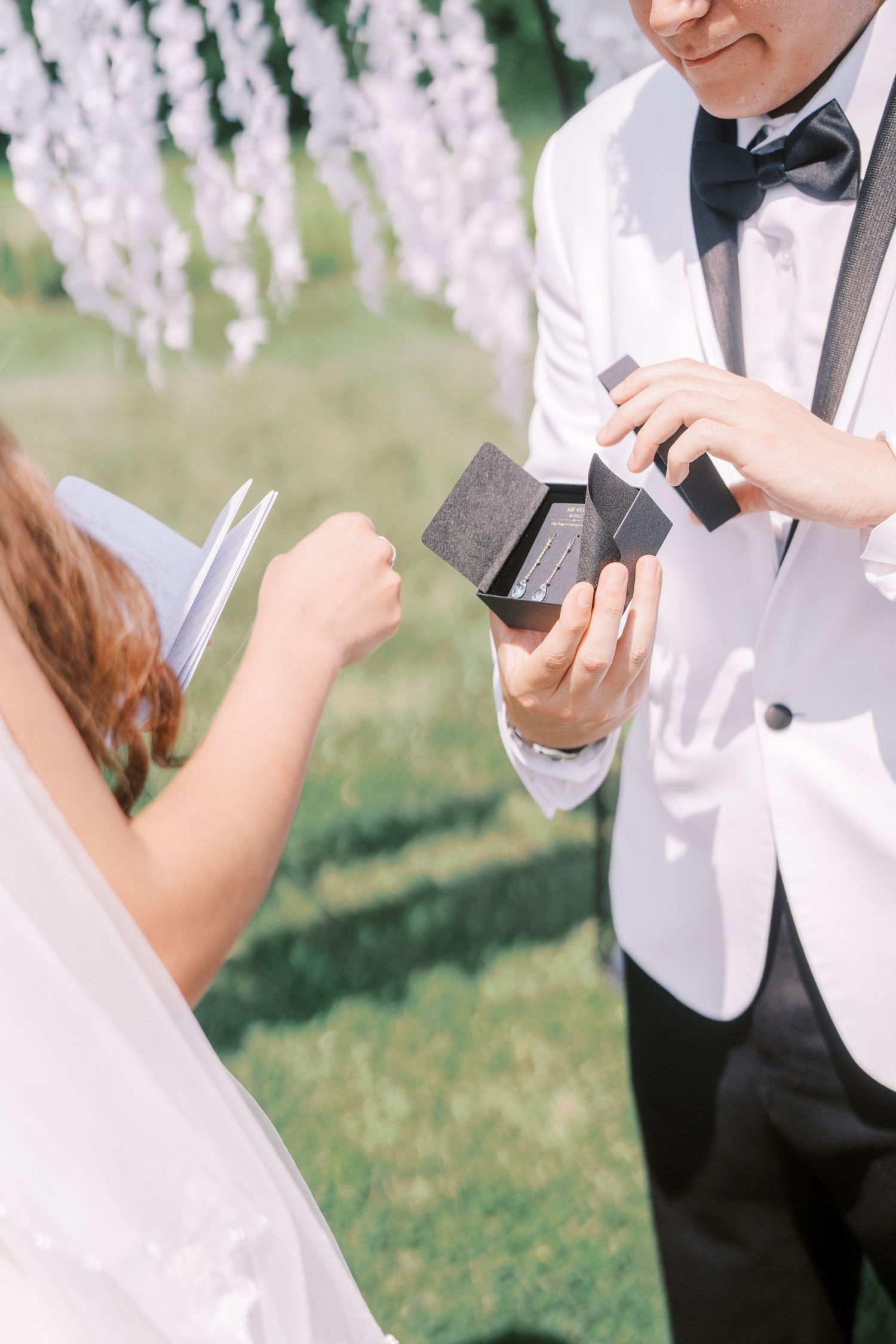 A couple standing outdoors exchanges notes and boxes during a wedding ceremony with white wisteria flowers in background.
