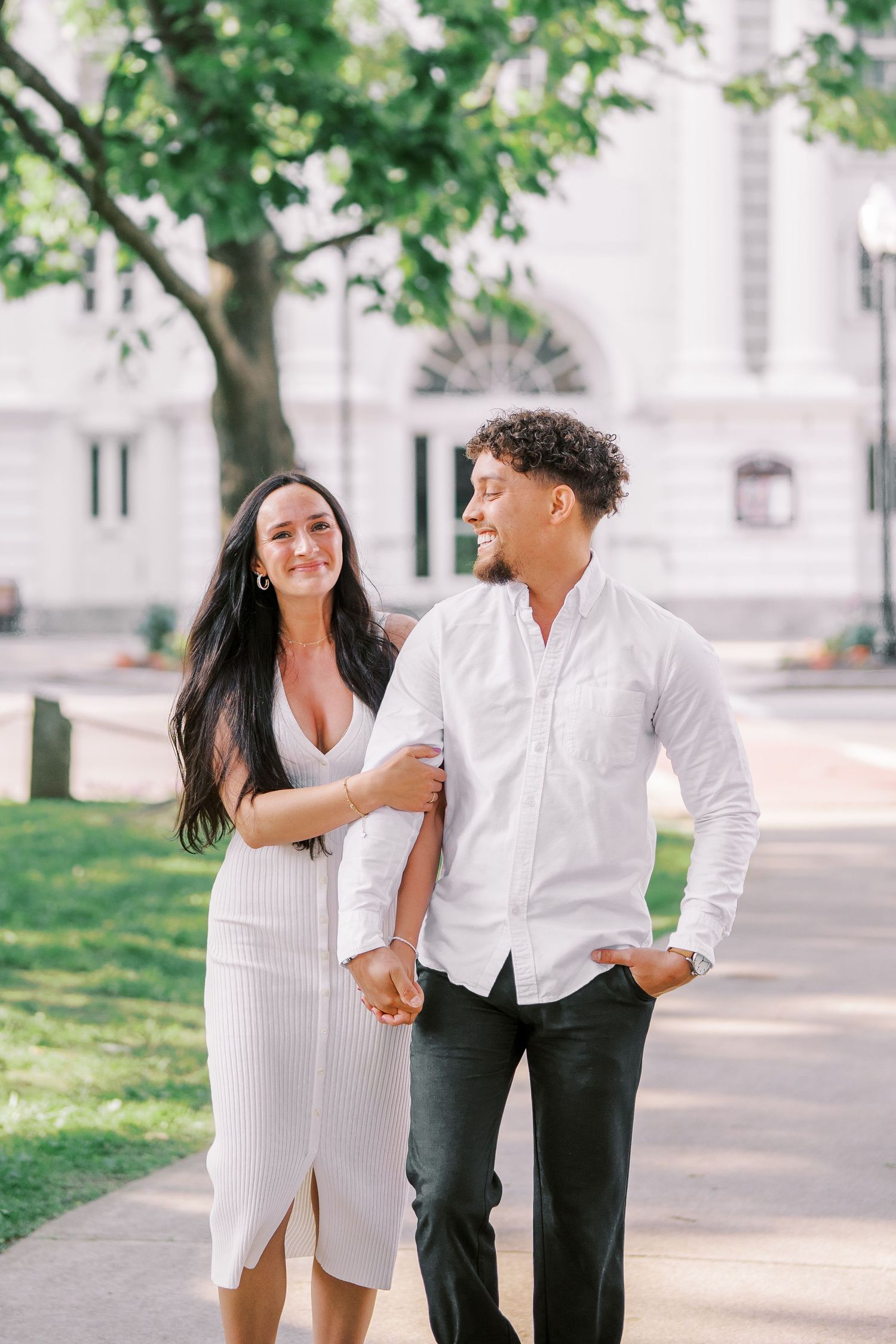 Couple dressed in white walking and laughing together on a tree-lined path during a sunny day.