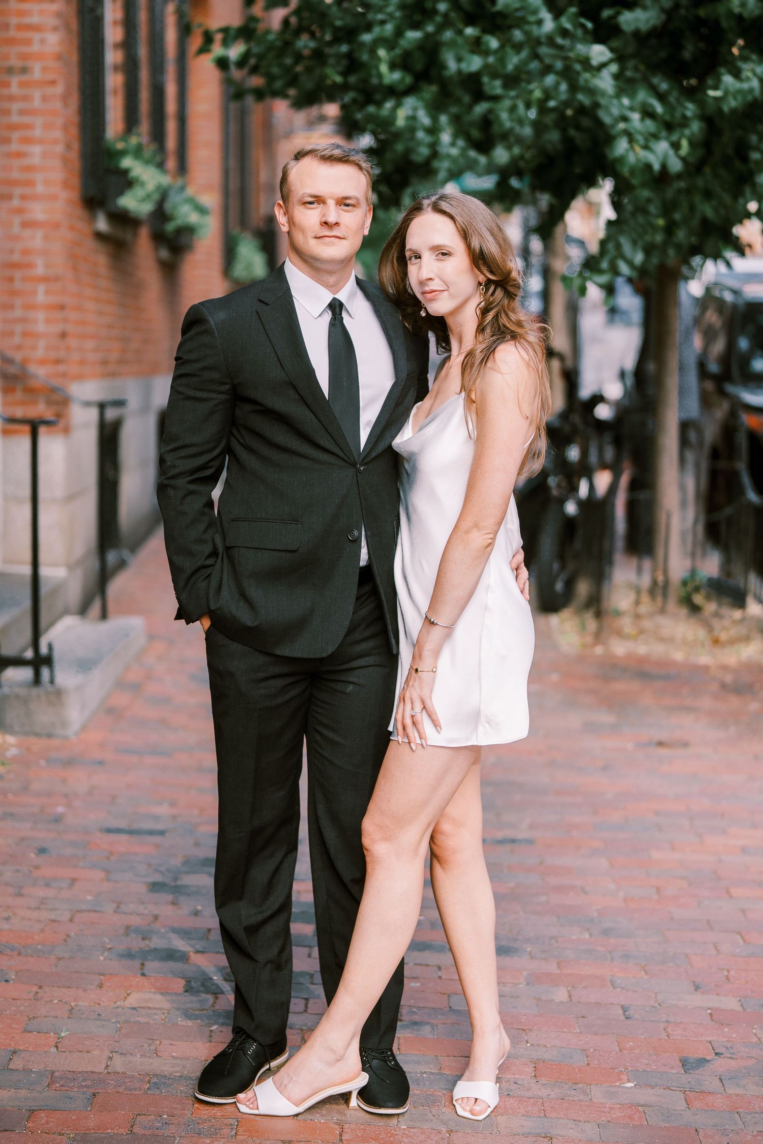 A couple poses intimately on a brick sidewalk with trees and historic buildings in the background.