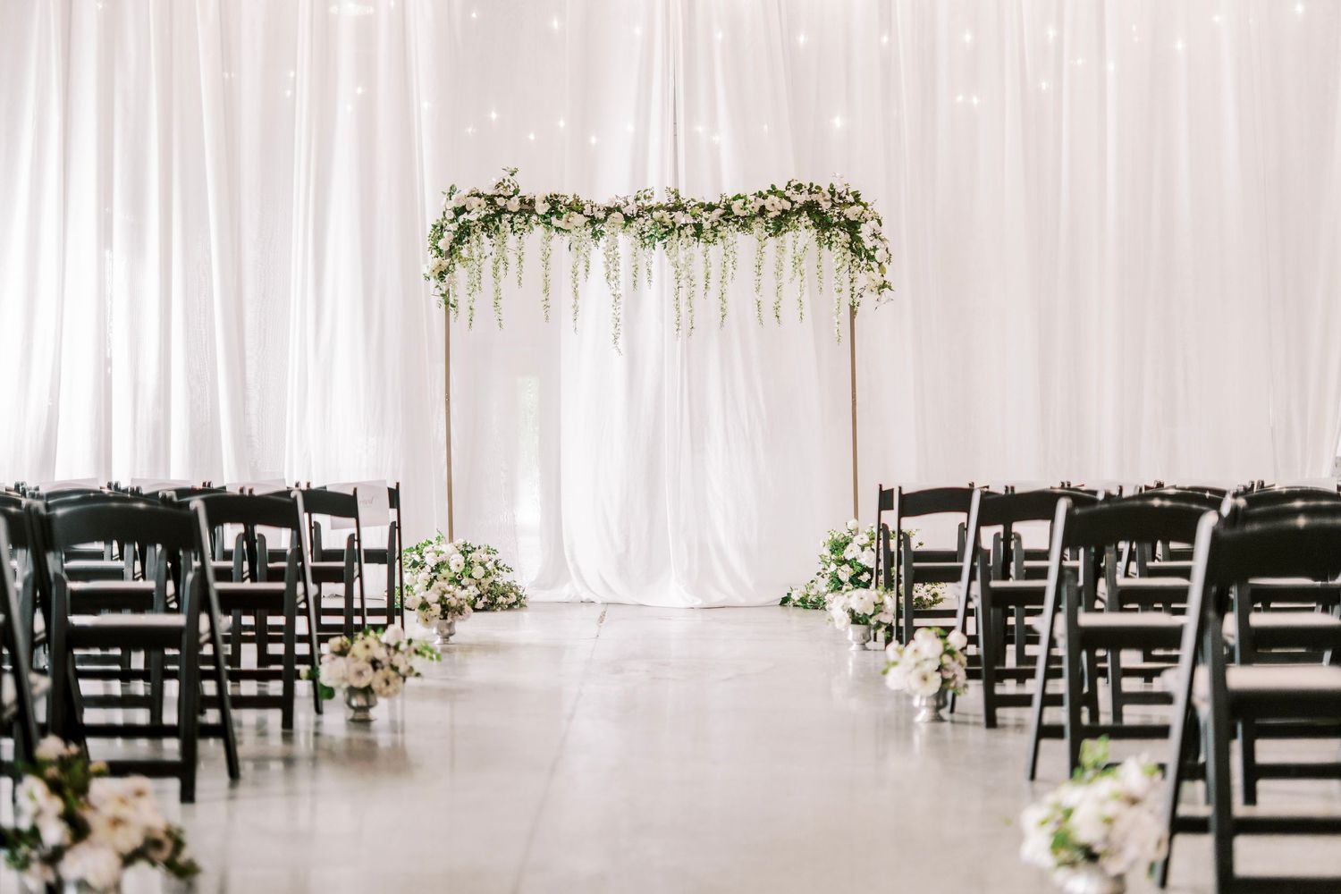 Minimalist wedding ceremony setup with white drapes, floral arch, and black chairs in rows along the aisle.