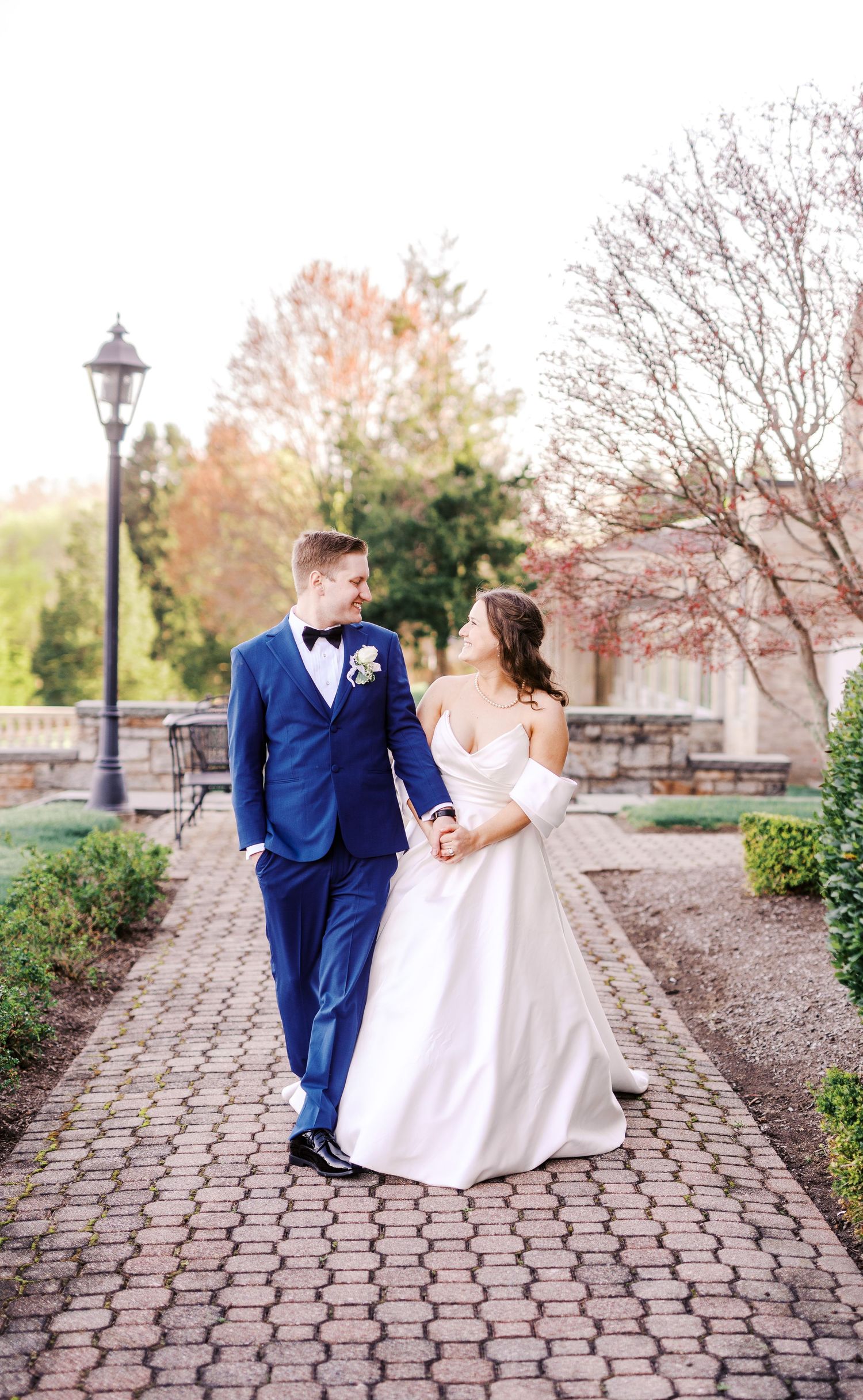 Newlyweds in blue suit and white dress walking together on brick pathway with spring blossoms.