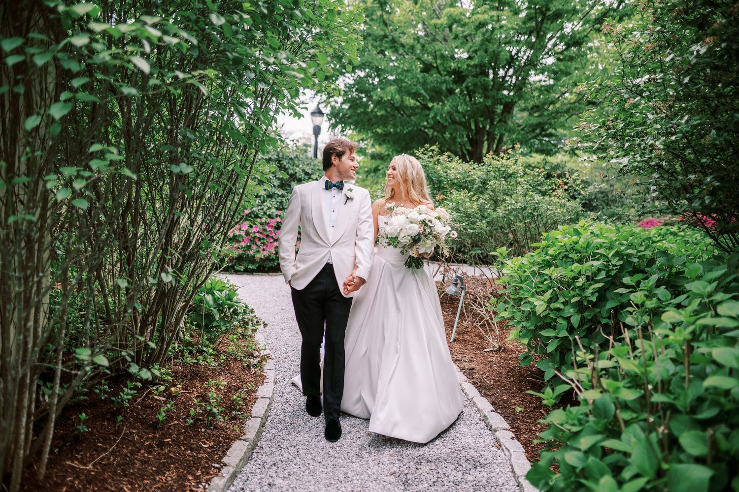A couple in wedding attire walks hand in hand down a lush green garden path surrounded by natural foliage.