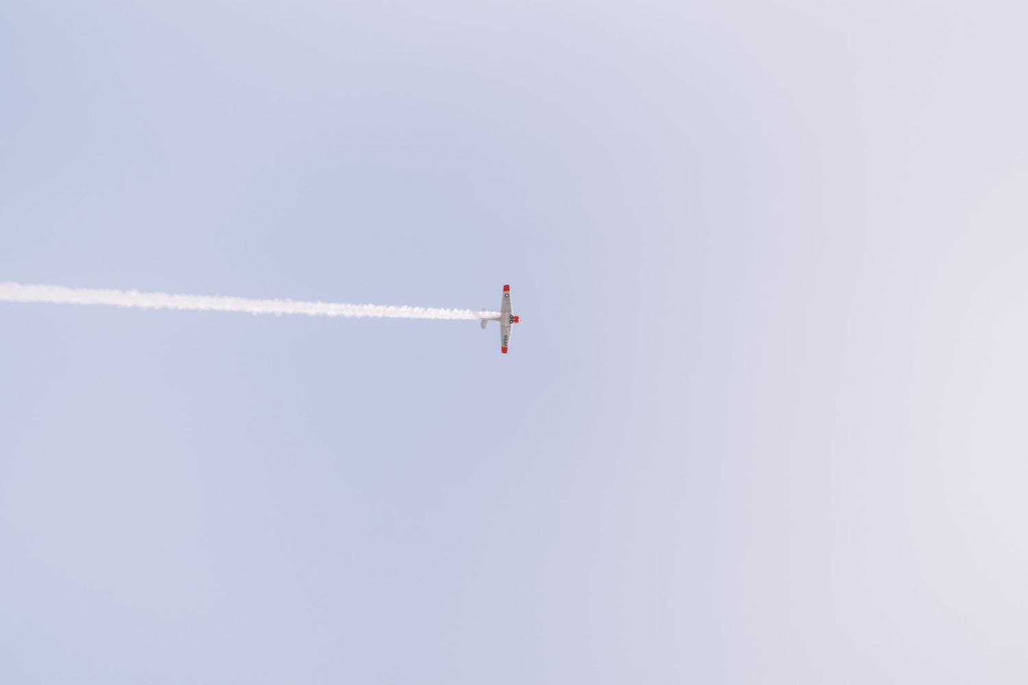 Small airplane flying high in the pale blue sky leaving a white contrail streak behind it.