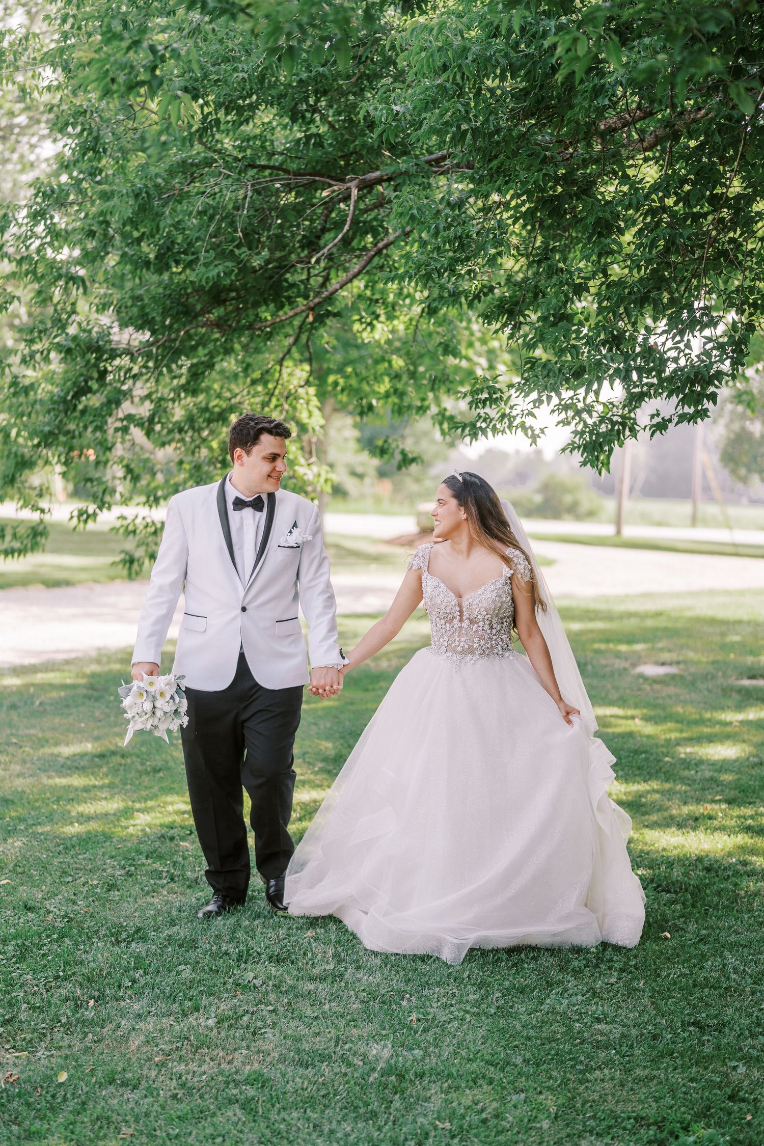 A couple in wedding attire walk together on green grass under a shady tree during an outdoor summer celebration.
