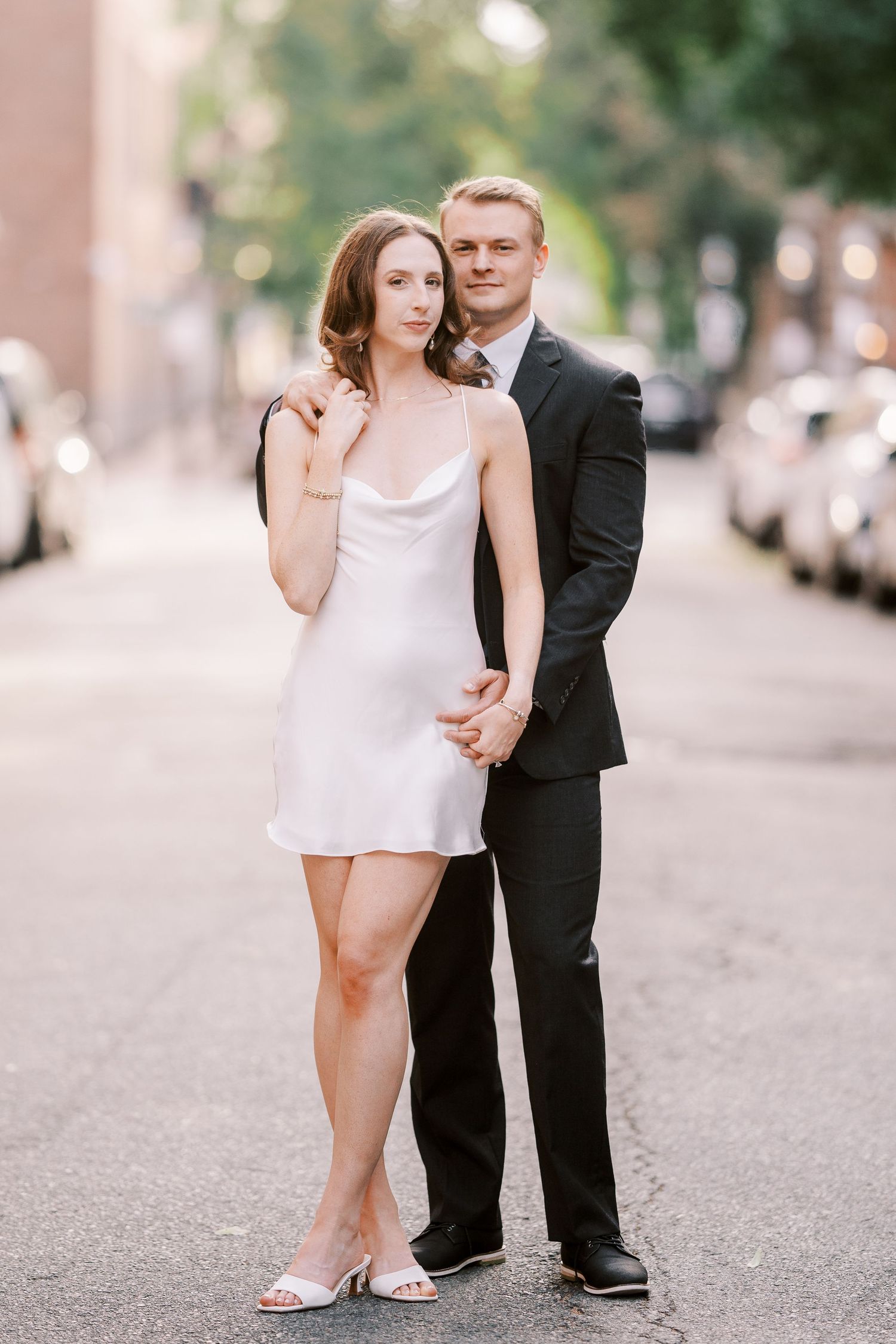 A couple poses together on a city street, with her in a white slip dress and him in a black suit.