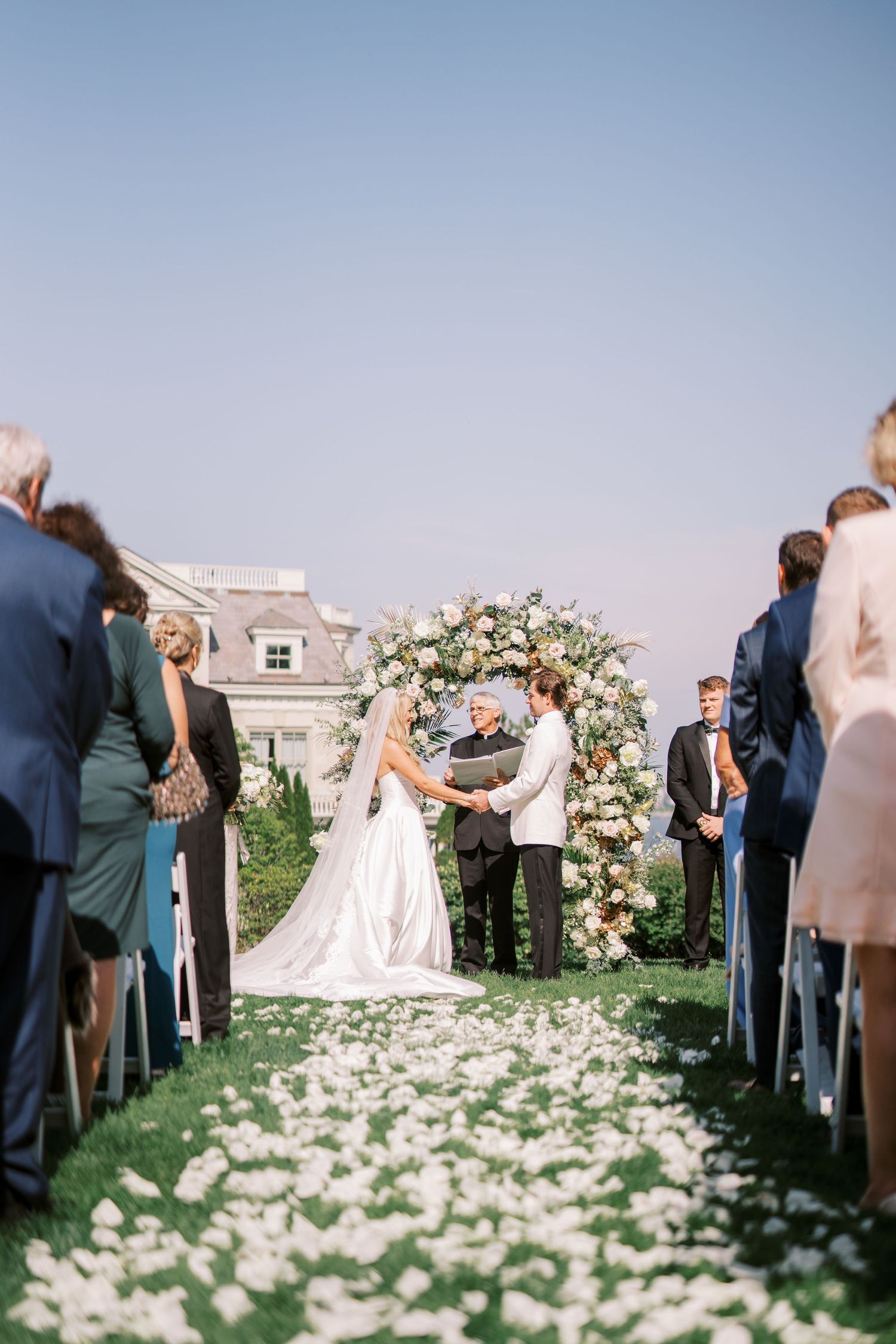 Elegant outdoor wedding ceremony with flower petal-lined aisle and floral arch against a clear blue sky.
