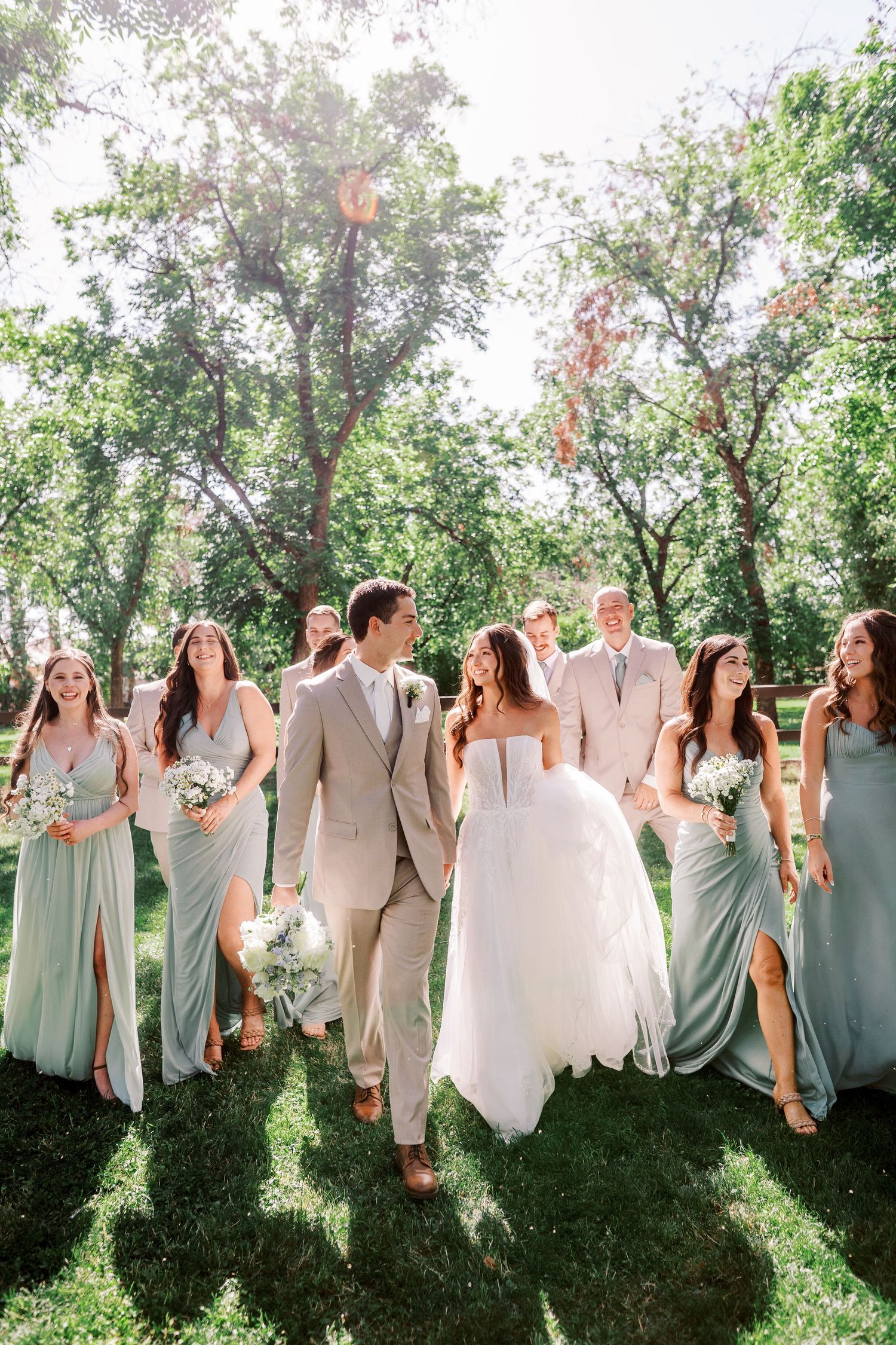Wedding party walks together outdoors in sage green bridesmaid dresses and beige suit on sunny summer day.