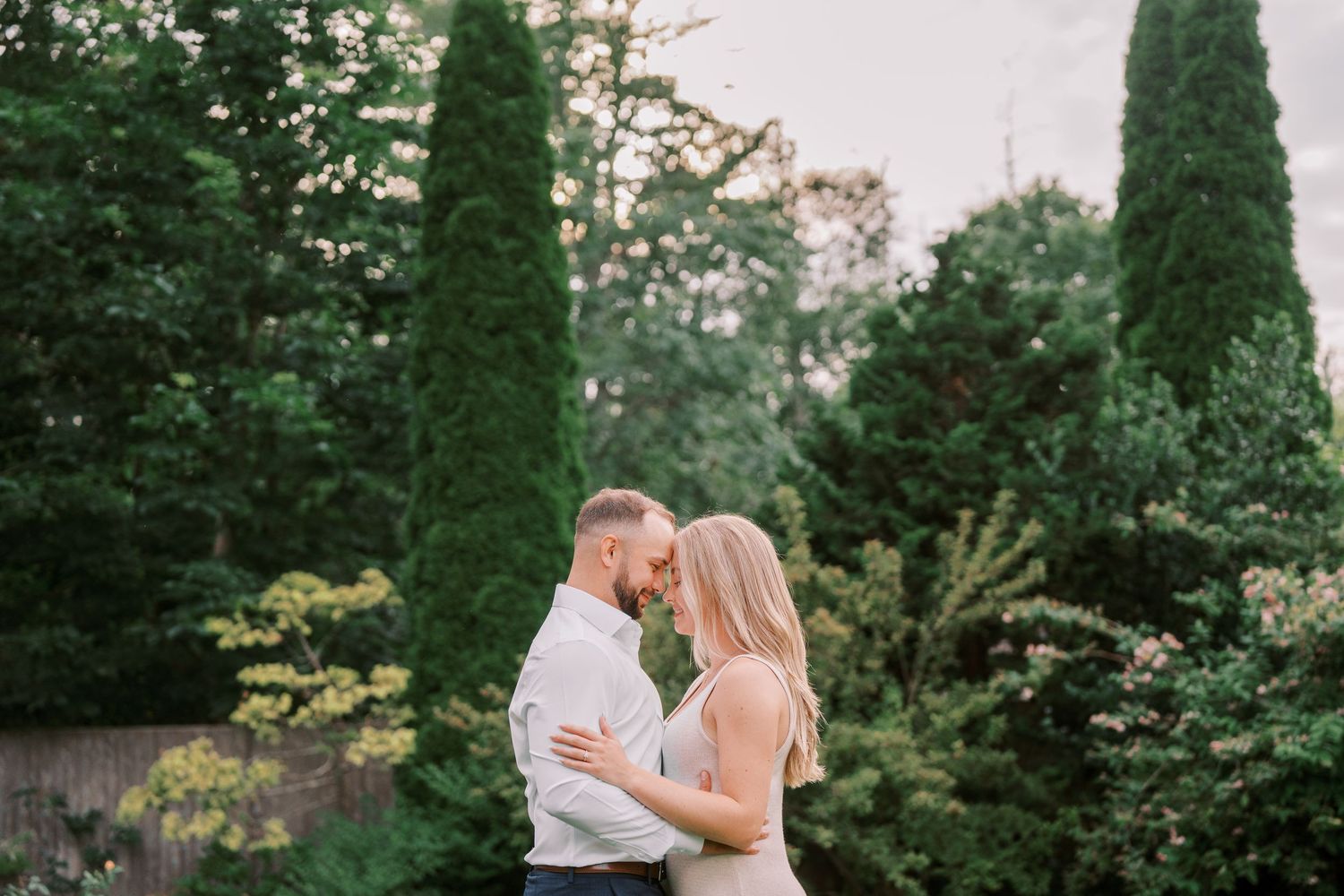 A romantic outdoor couple embraces surrounded by lush greenery and tall cypress trees at sunset.