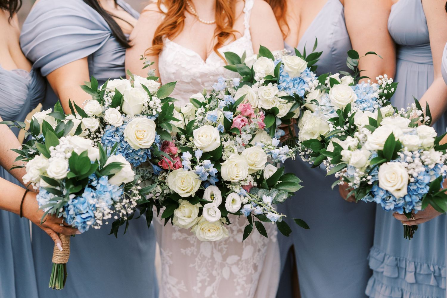 Wedding bouquets featuring white roses, blue hydrangeas and green foliage held against dusty blue bridesmaid dresses.