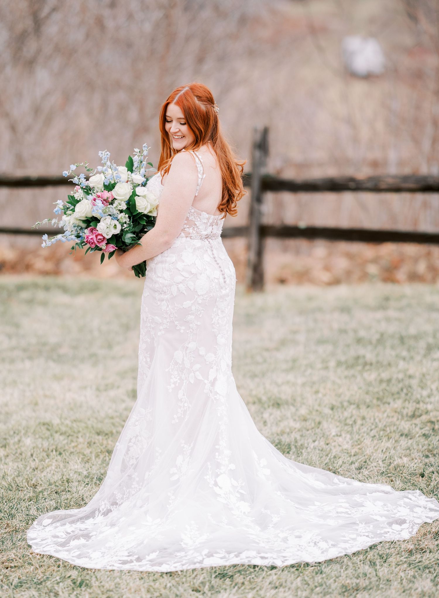 Bride in lace wedding dress with long train holding pink and white floral bouquet in outdoor setting.