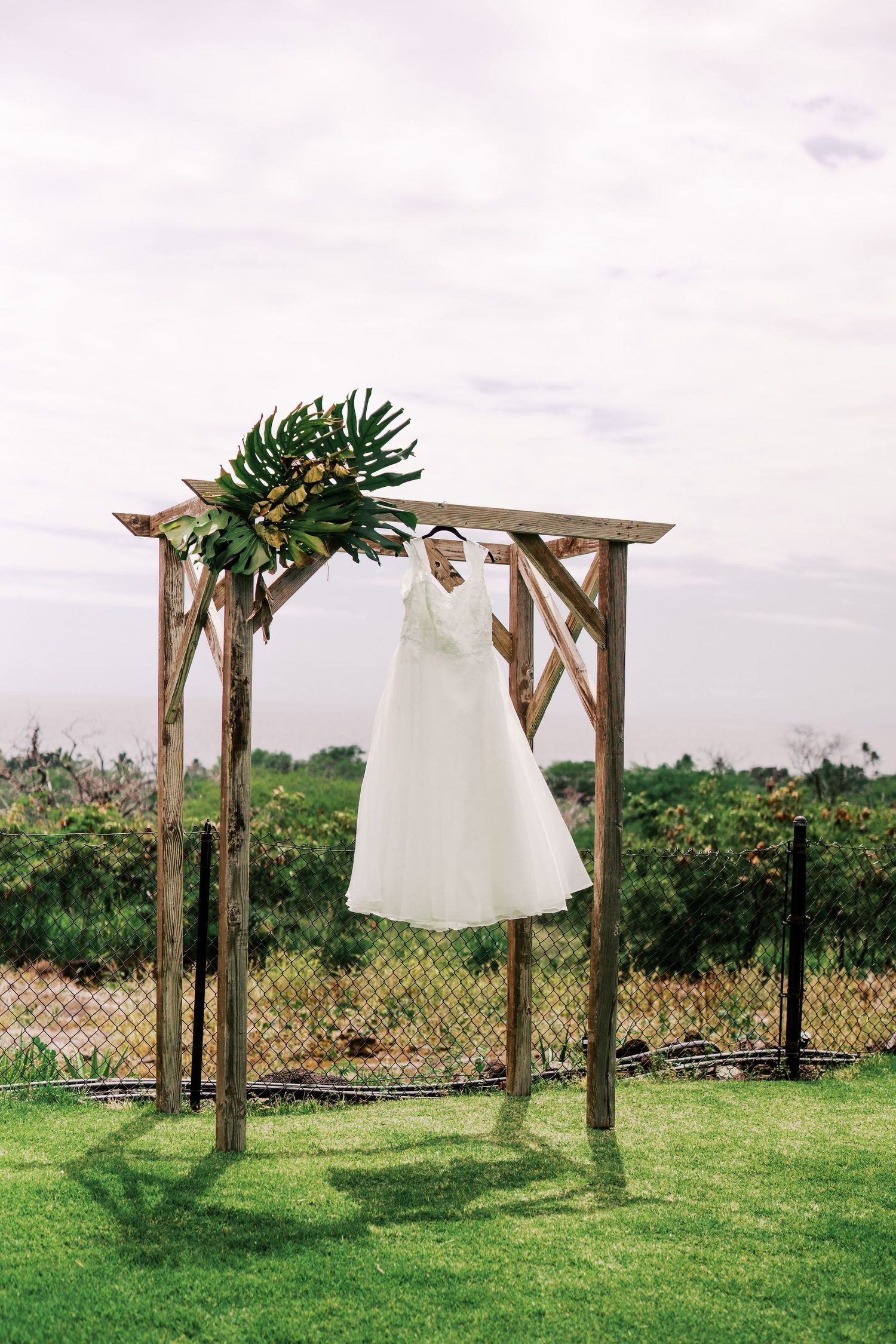 White wedding dress hanging on rustic wooden arch decorated with greenery in outdoor garden setting.