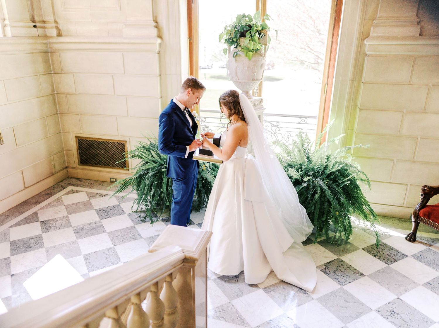 A couple exchanging rings during an intimate wedding ceremony in a bright sunlit venue with checkered marble floors and ferns.