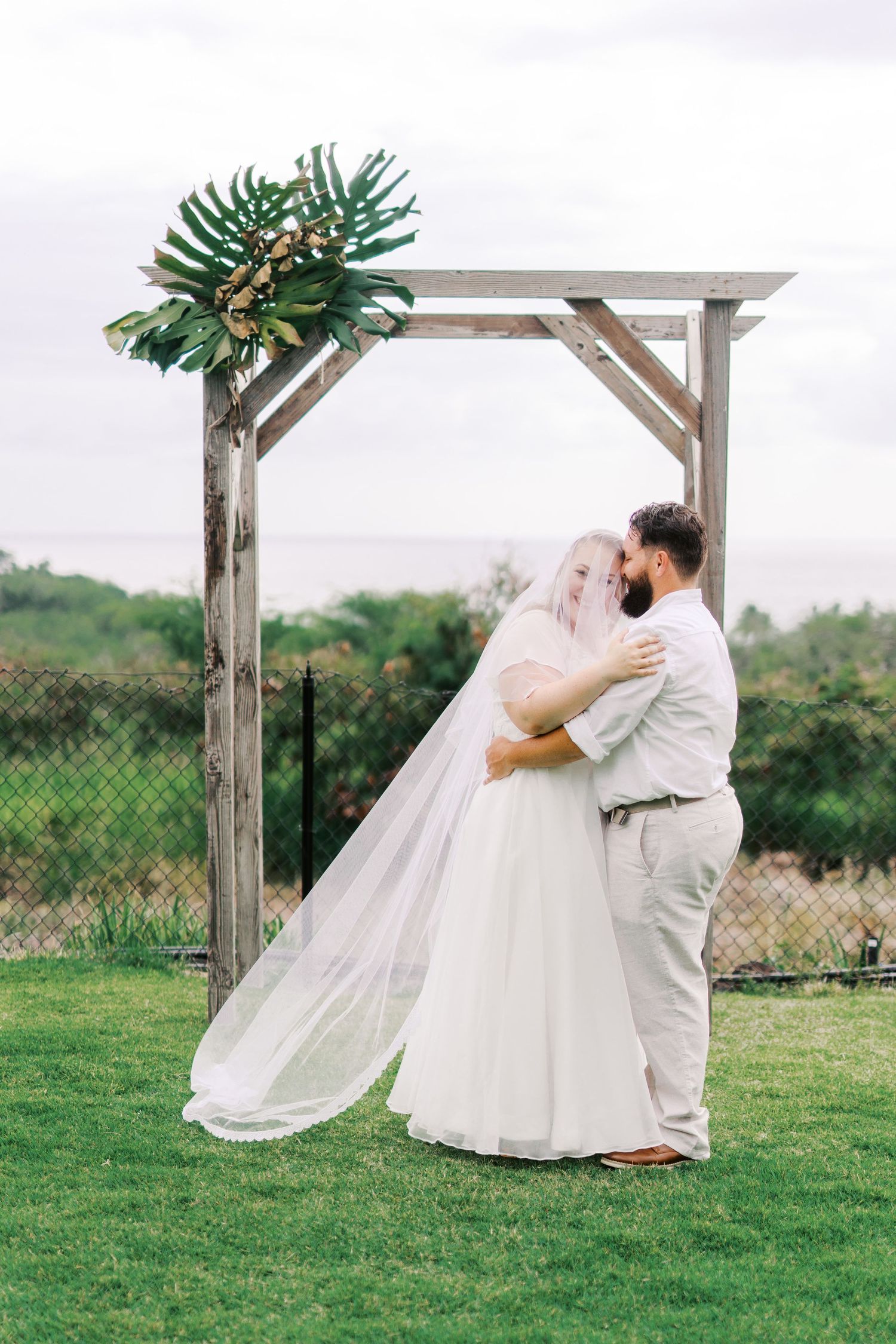 A wedding couple embraces under a rustic wooden arch adorned with tropical greenery.
