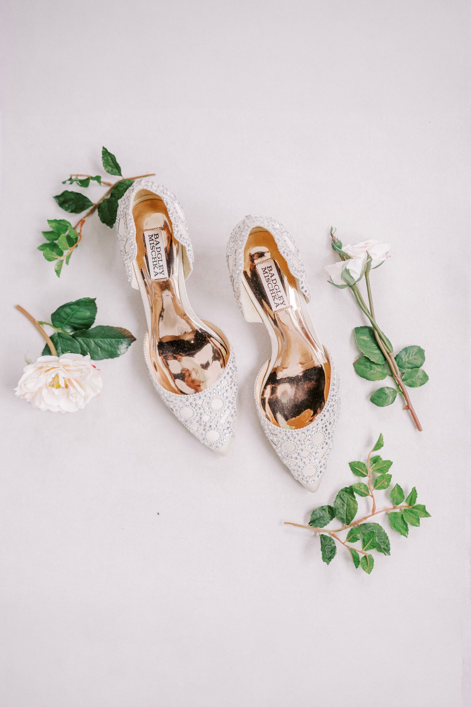 White glitter bridal slingback heels arranged with delicate pink roses and eucalyptus leaves on light gray background.