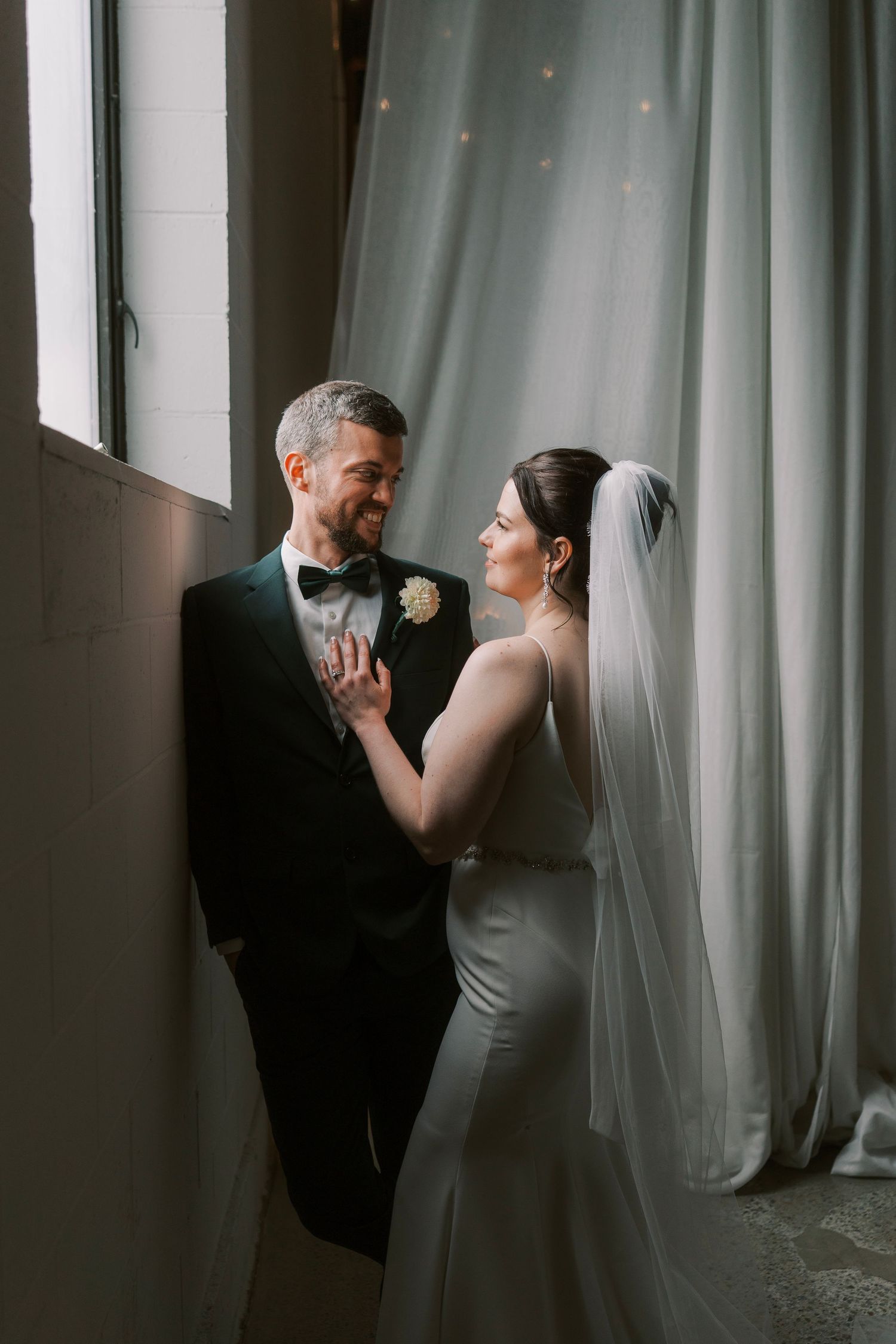A tender moment between newlyweds near a window with soft lighting and flowing curtains.