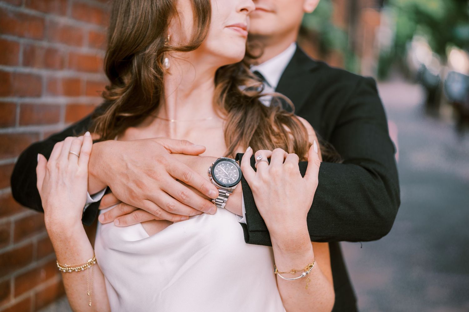 A closeup of embracing couple showing hands and jewelry against a brick wall backdrop.