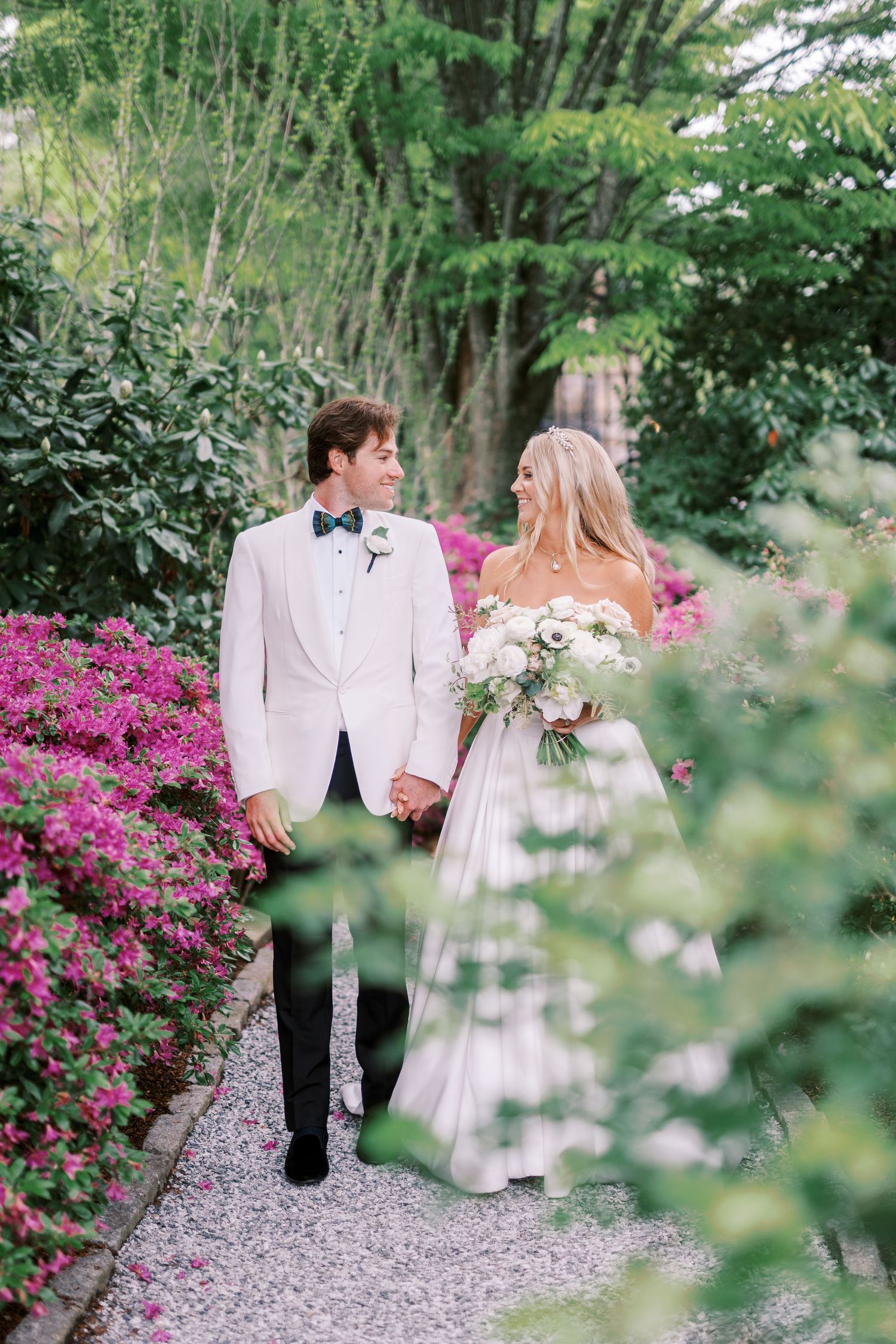 A couple walks along a garden path lined with vibrant pink azalea bushes and lush greenery during their wedding day.