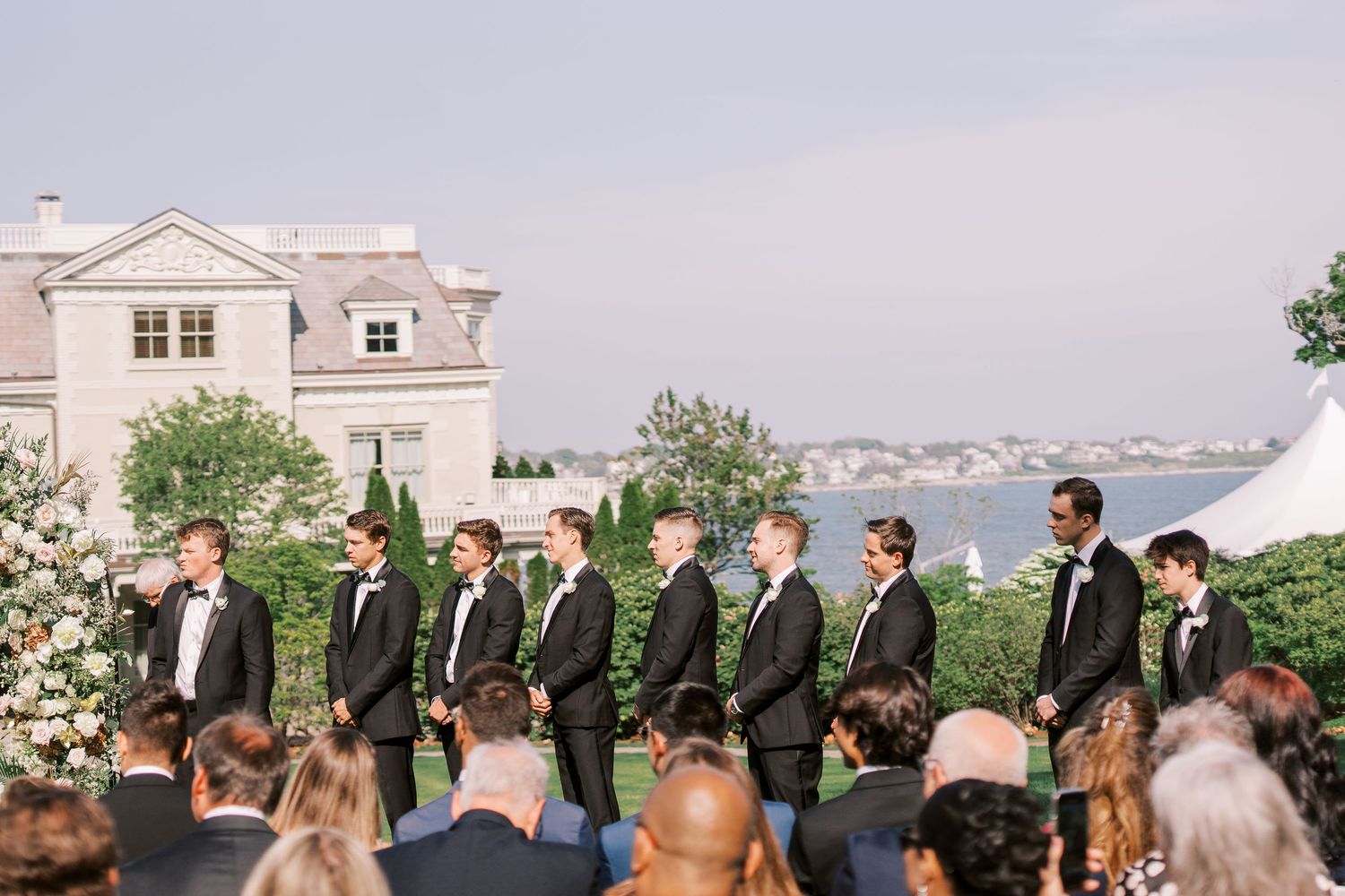 Elegant outdoor wedding ceremony overlooking the water with groomsmen in dark suits lined up at the altar.