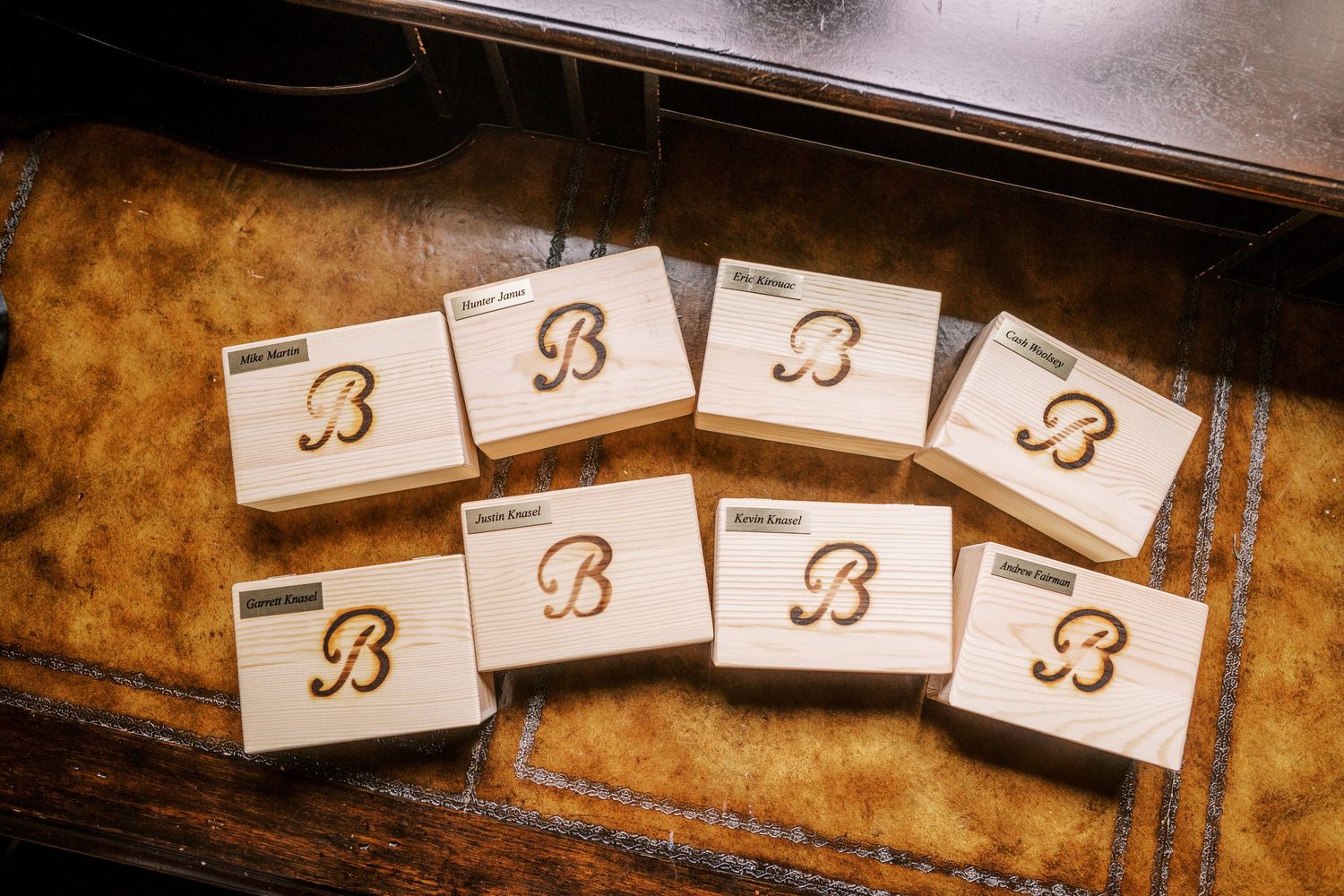 Eight wooden matchboxes with an ornate letter B engraved or burnt into their surfaces arranged in a curved pattern.