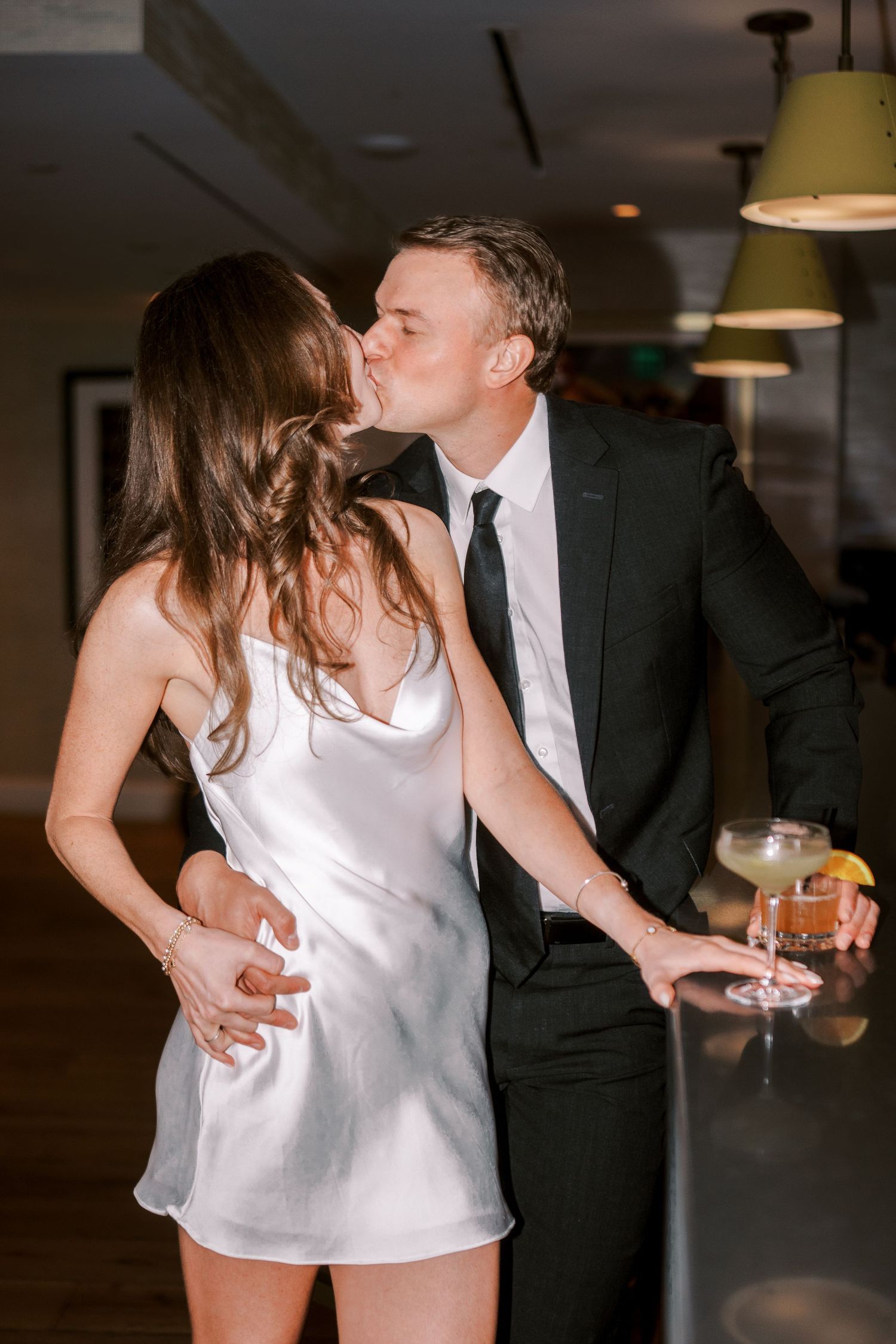 A romantic couple share an intimate moment at a dimly lit bar, with her in a white dress and him in a dark suit.