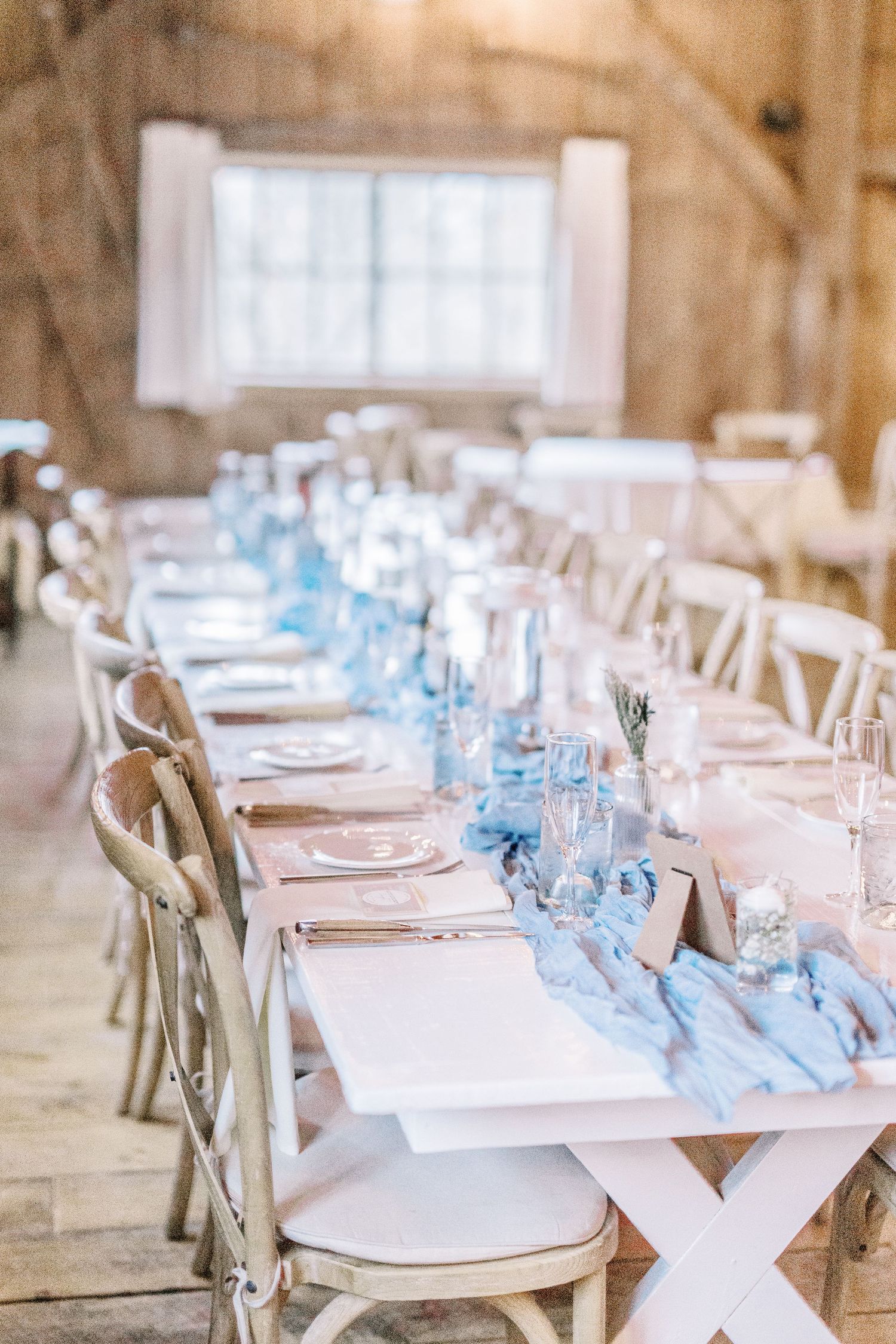 Long rustic wedding table with white chairs and light blue runner in a barn setting.