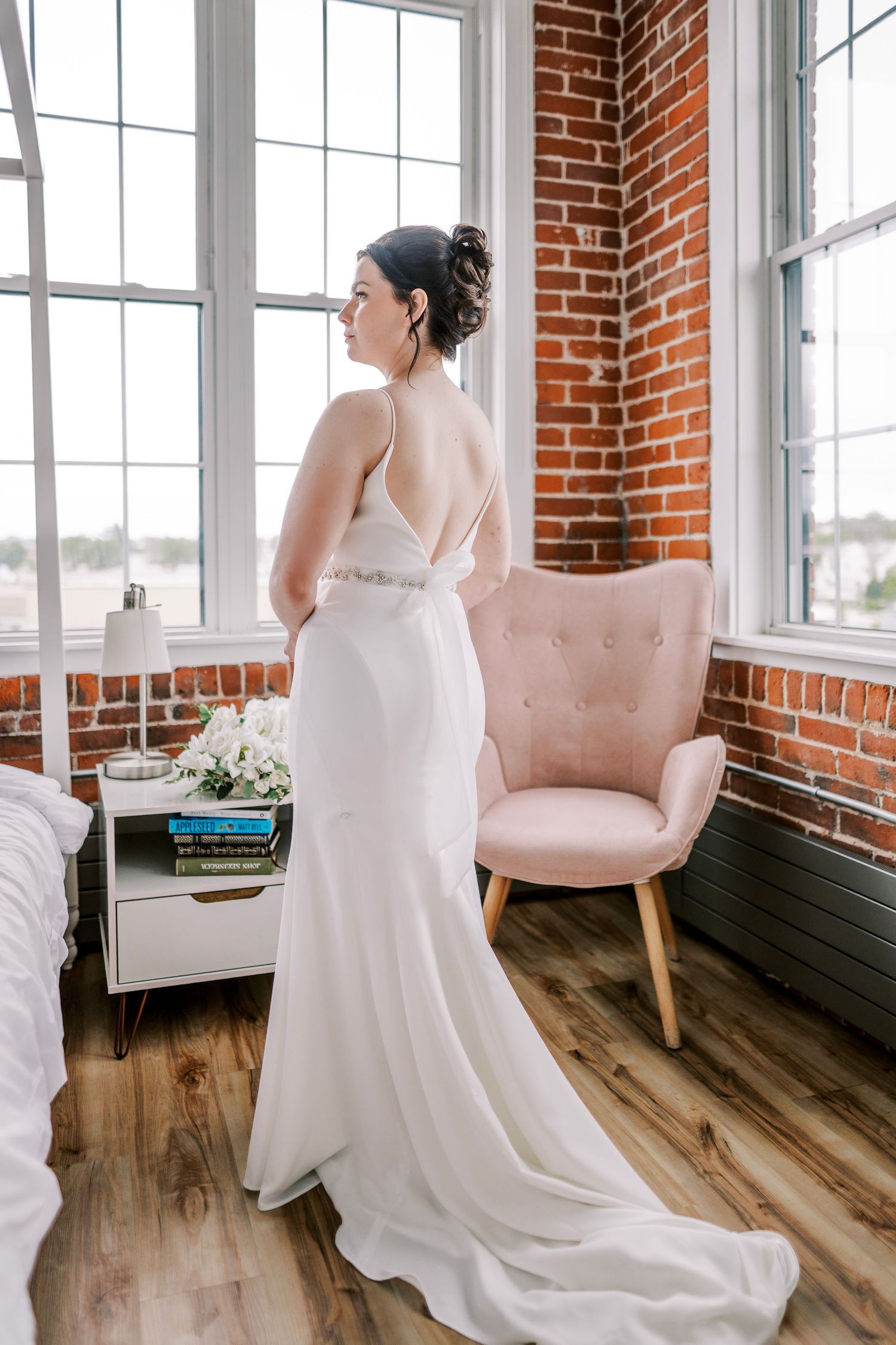Elegant white wedding dress with open back detail against exposed brick wall and large windows.