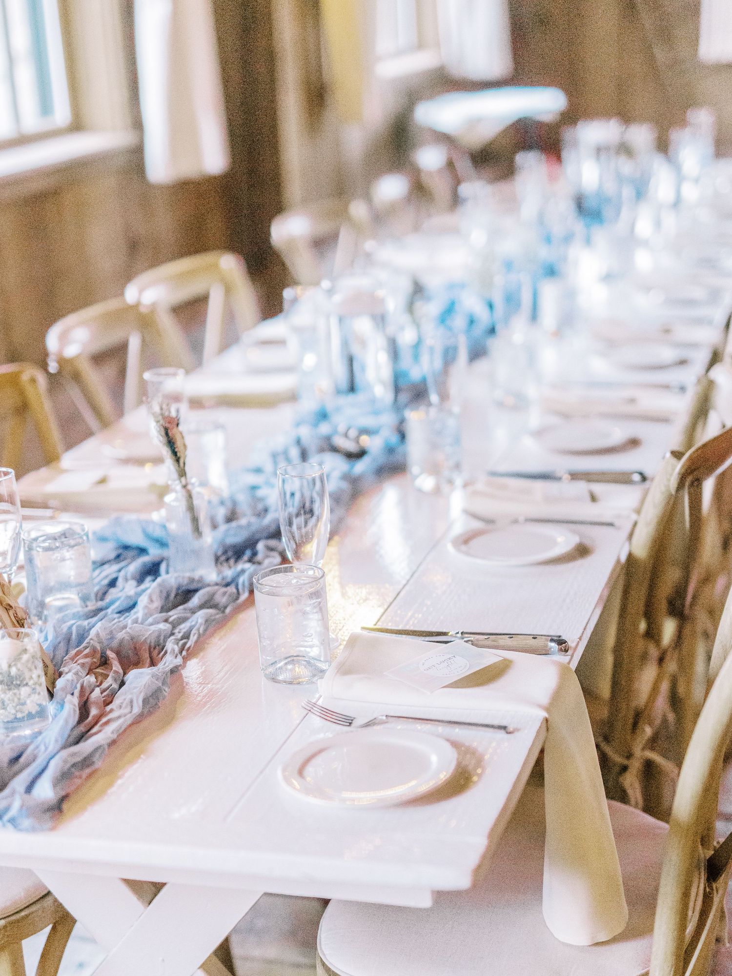 Long elegant wedding reception table with blue runner, white place settings and wooden crossback chairs.