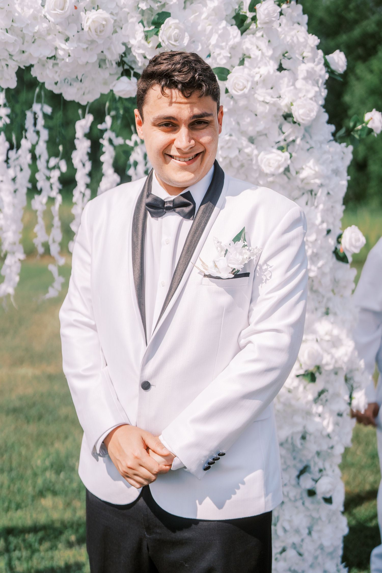 A person in a white tuxedo jacket and bowtie stands next to white floral wedding decorations outdoors.