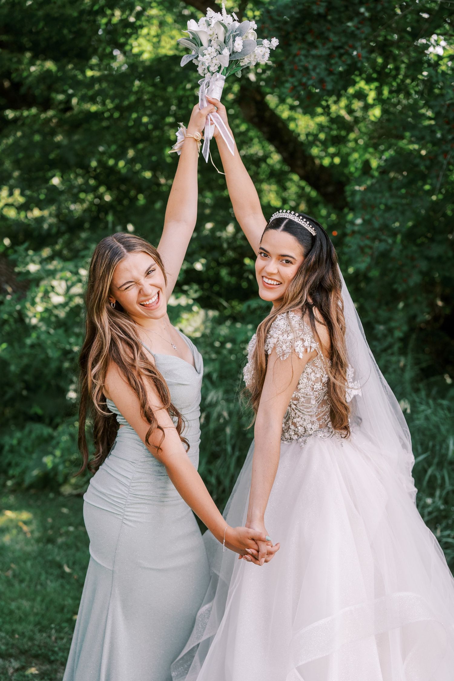 Bridesmaids and bride dancing and laughing together in an outdoor garden setting wearing grey and white dresses.