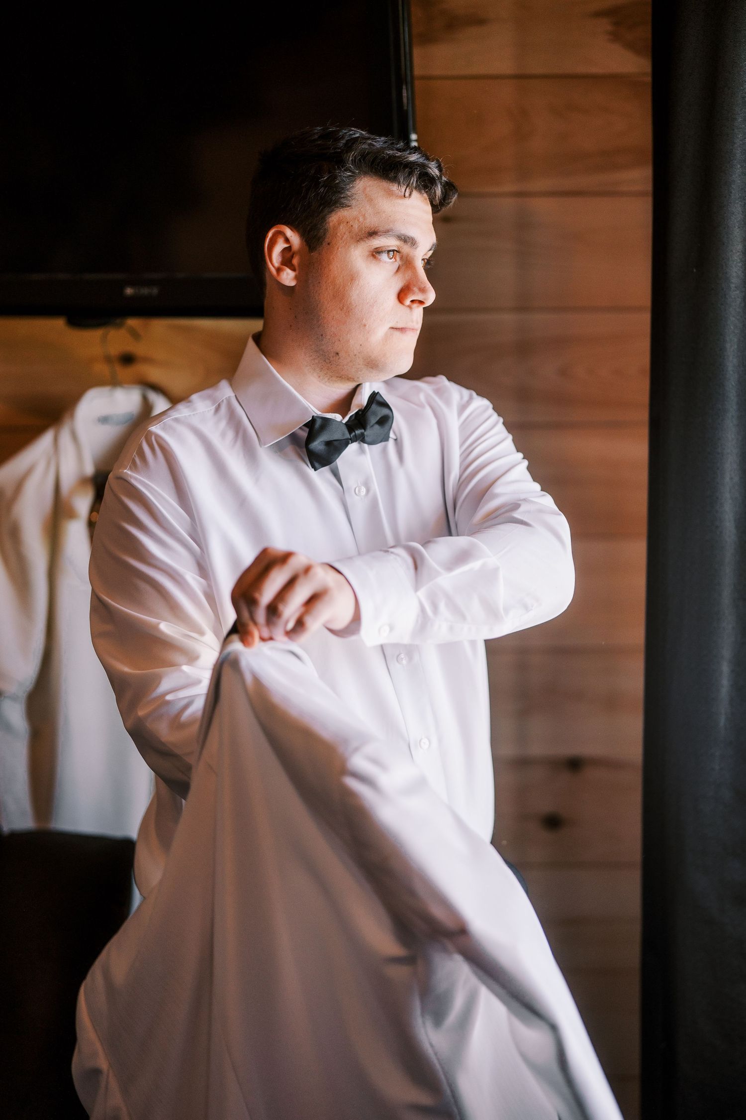 A person in a white dress shirt adjusts a black bow tie while getting ready in an interior with wood paneled walls.