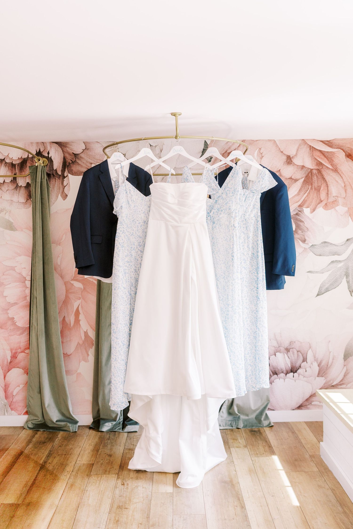A white wedding dress hangs on display with navy suits against a pink floral wallpapered wall.