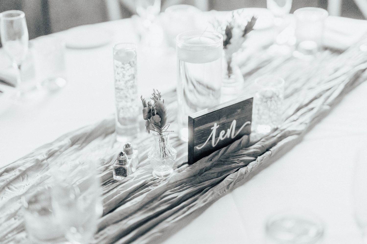 A high-key black and white table setting features a glass jar with a handwritten label amidst delicate linens.