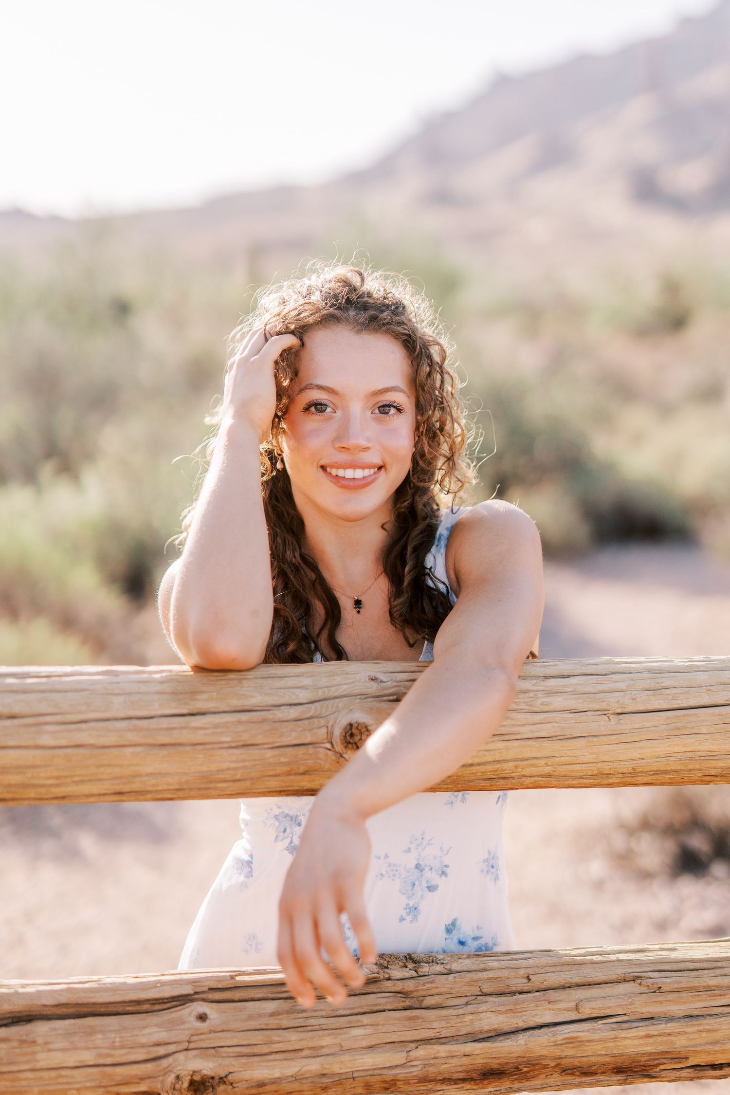 A sunny desert portrait shows someone peeking playfully over a wooden log fence with mountains visible in the background.